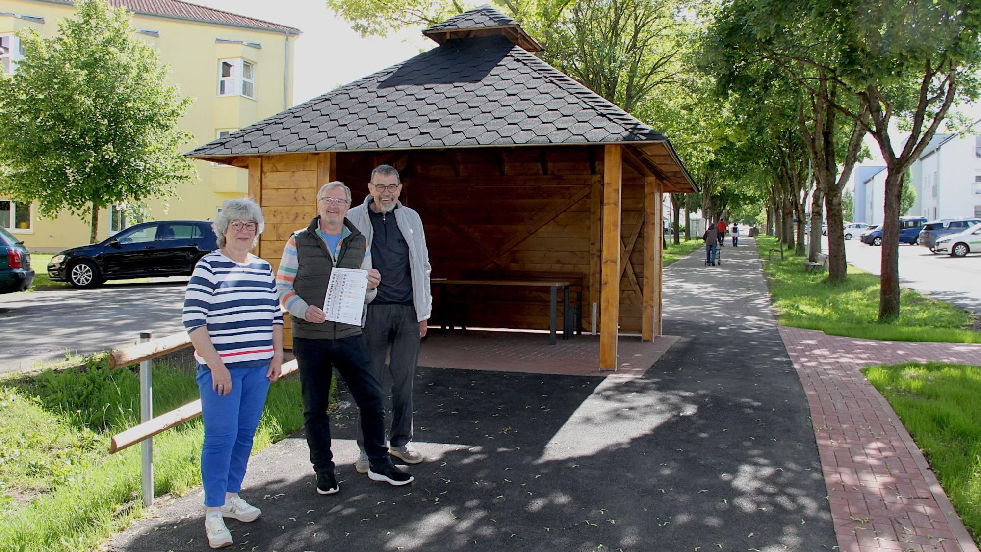 Eine Frau und zwei Männer stehen vor einem Pavillion, rundherum sind Bäume und Häuser zu erkennen.