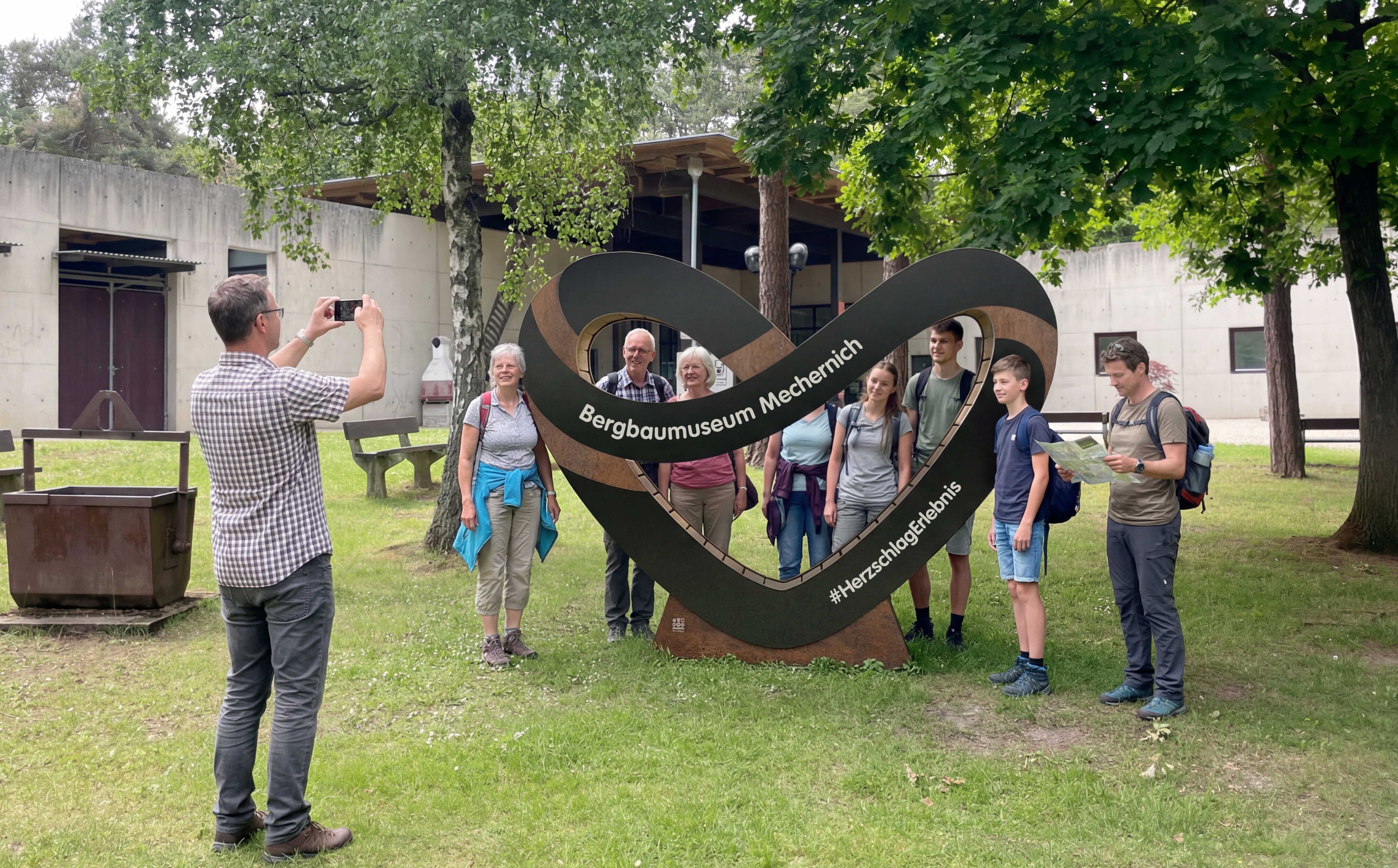 Menschen stehen um ein herförmiges Schild vor dem Bergbaumuseum in Mechernich und werden fotografiert.