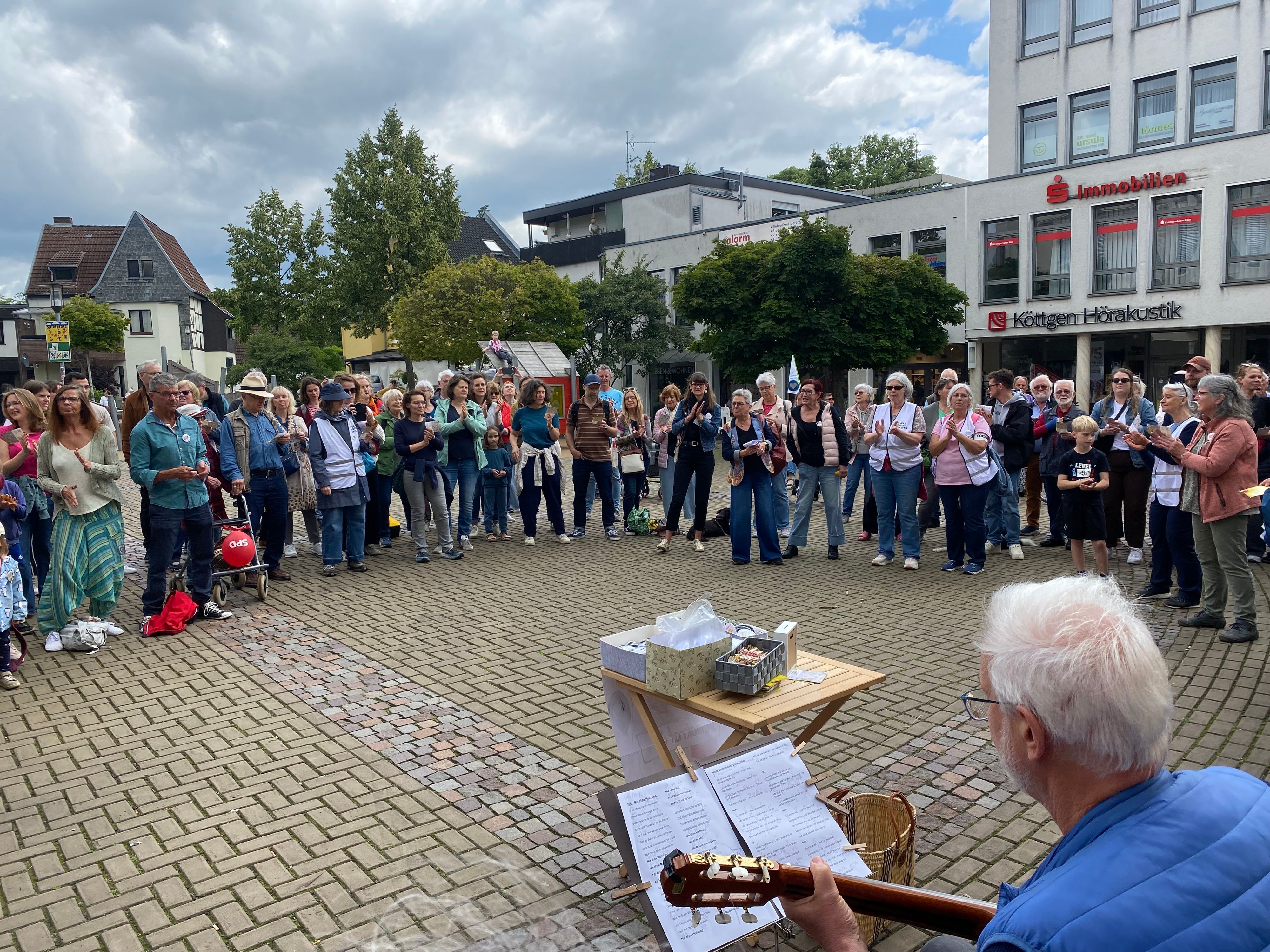 Zu sehen sind Menschen, die sich im Halbkreis aufgestellt haben. Im Vordergrund sitzt ein Mann mit einer Gitarre.