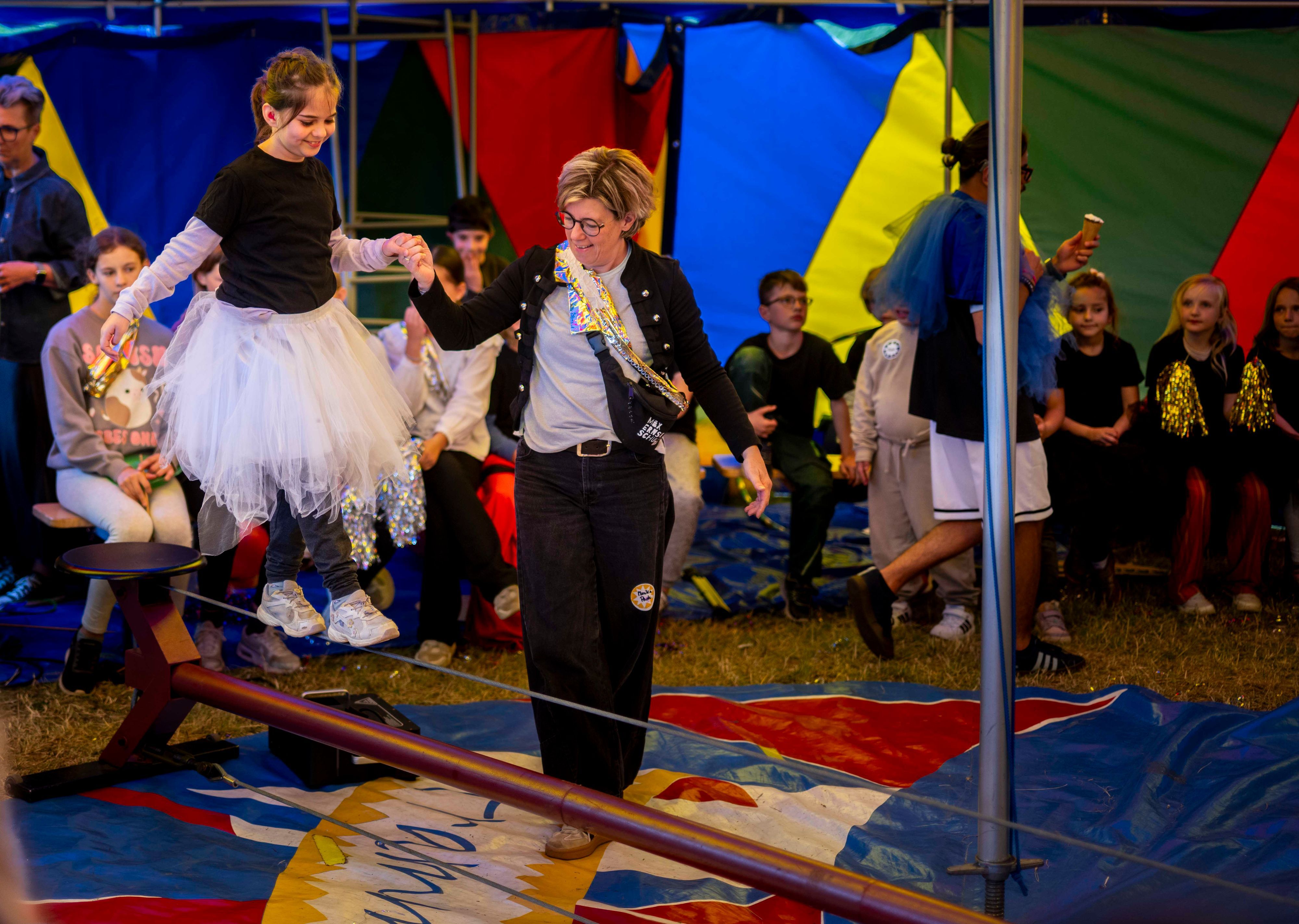 Ein Mädchen mit weißem Tutu balanciert an der Hand einer Frau über ein Drahtseil.