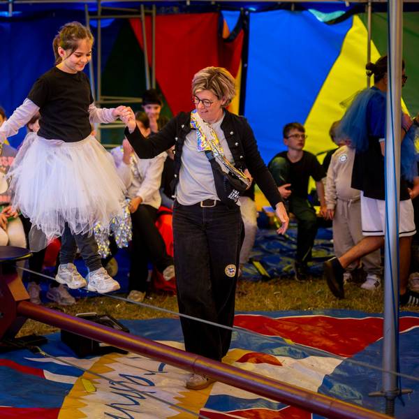 Ein Mädchen mit weißem Tutu balanciert an der Hand einer Frau über ein Drahtseil.