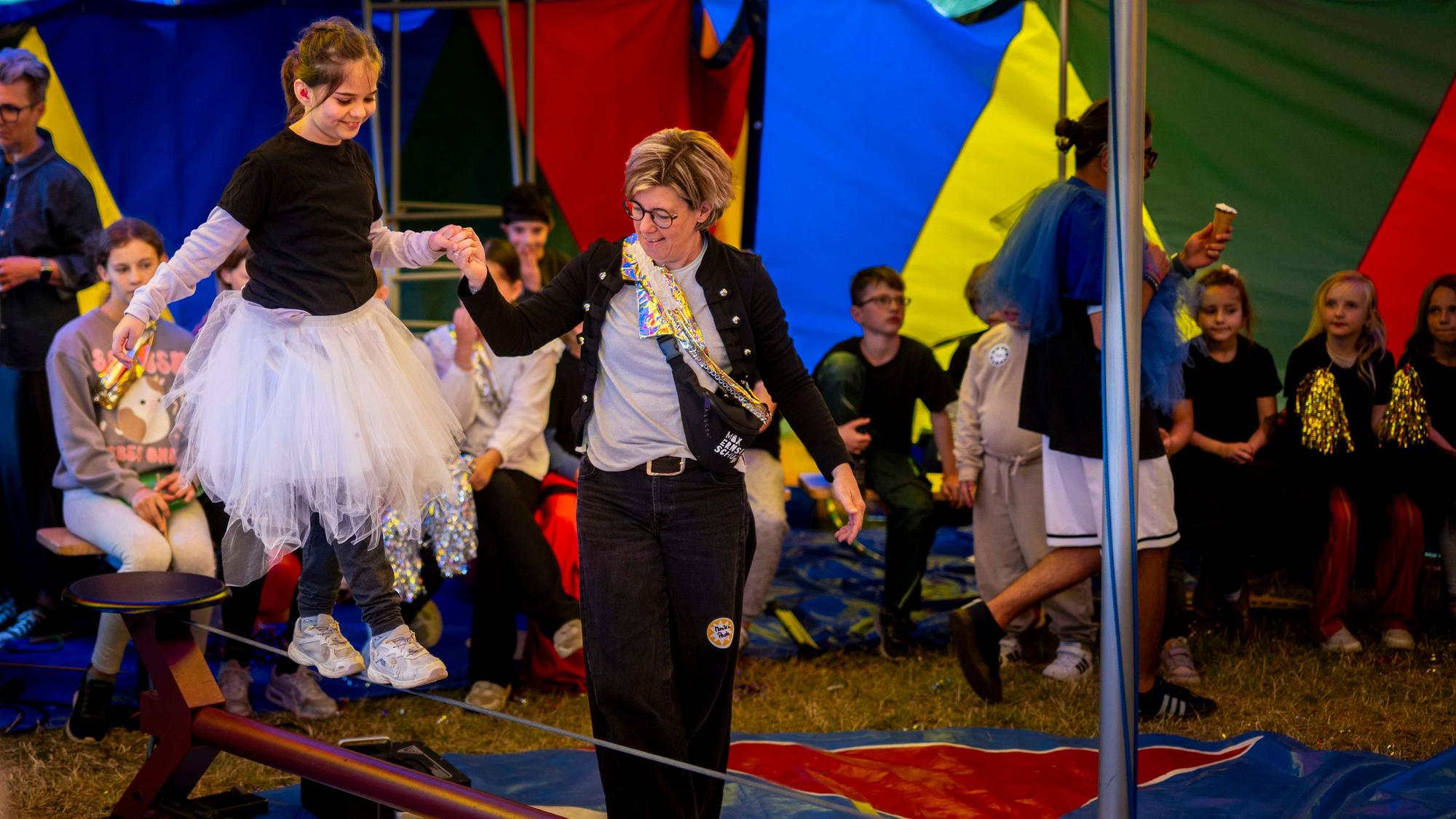 Ein Mädchen mit weißem Tutu balanciert an der Hand einer Frau über ein Drahtseil.