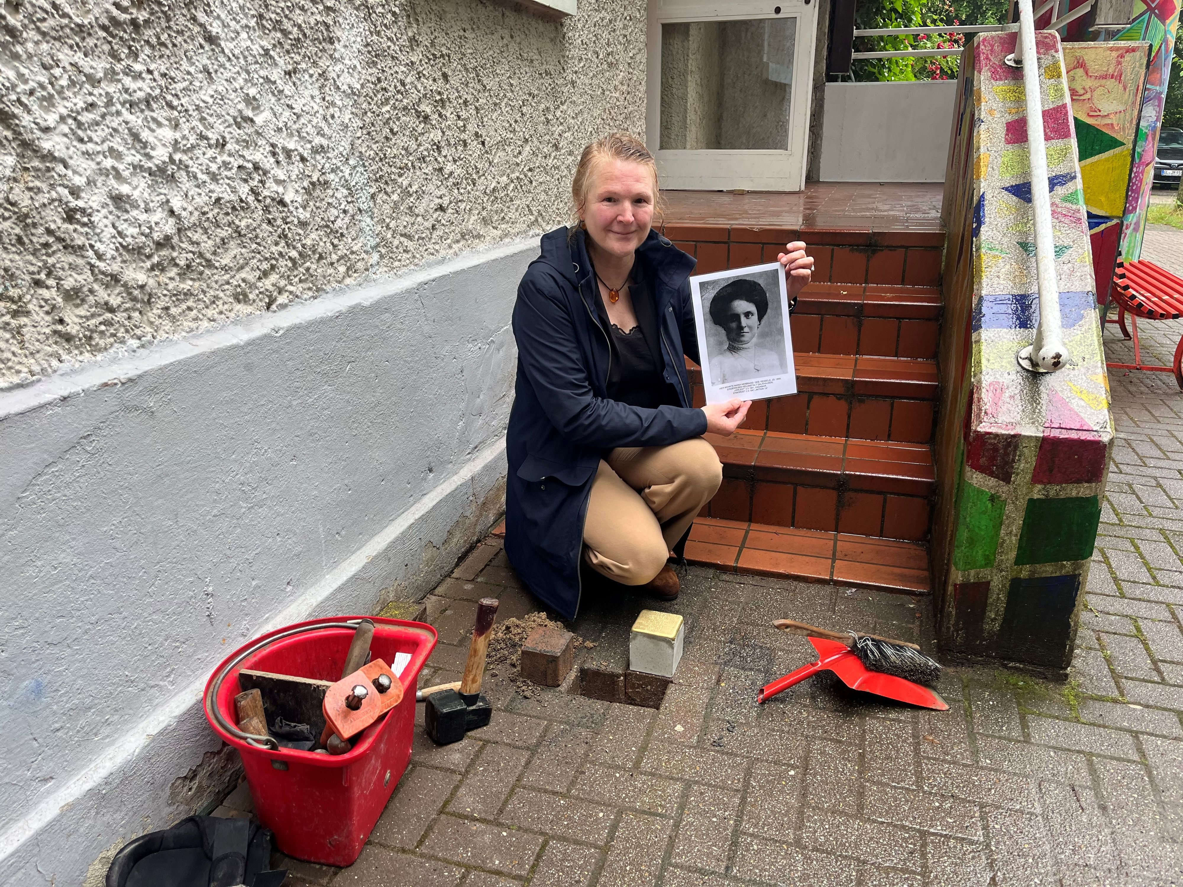 Eine Frau hockt mit einem schwarz-weißen Foto einer Frau vor einem Loch, in dem der Stolperstein verlegt werden soll.