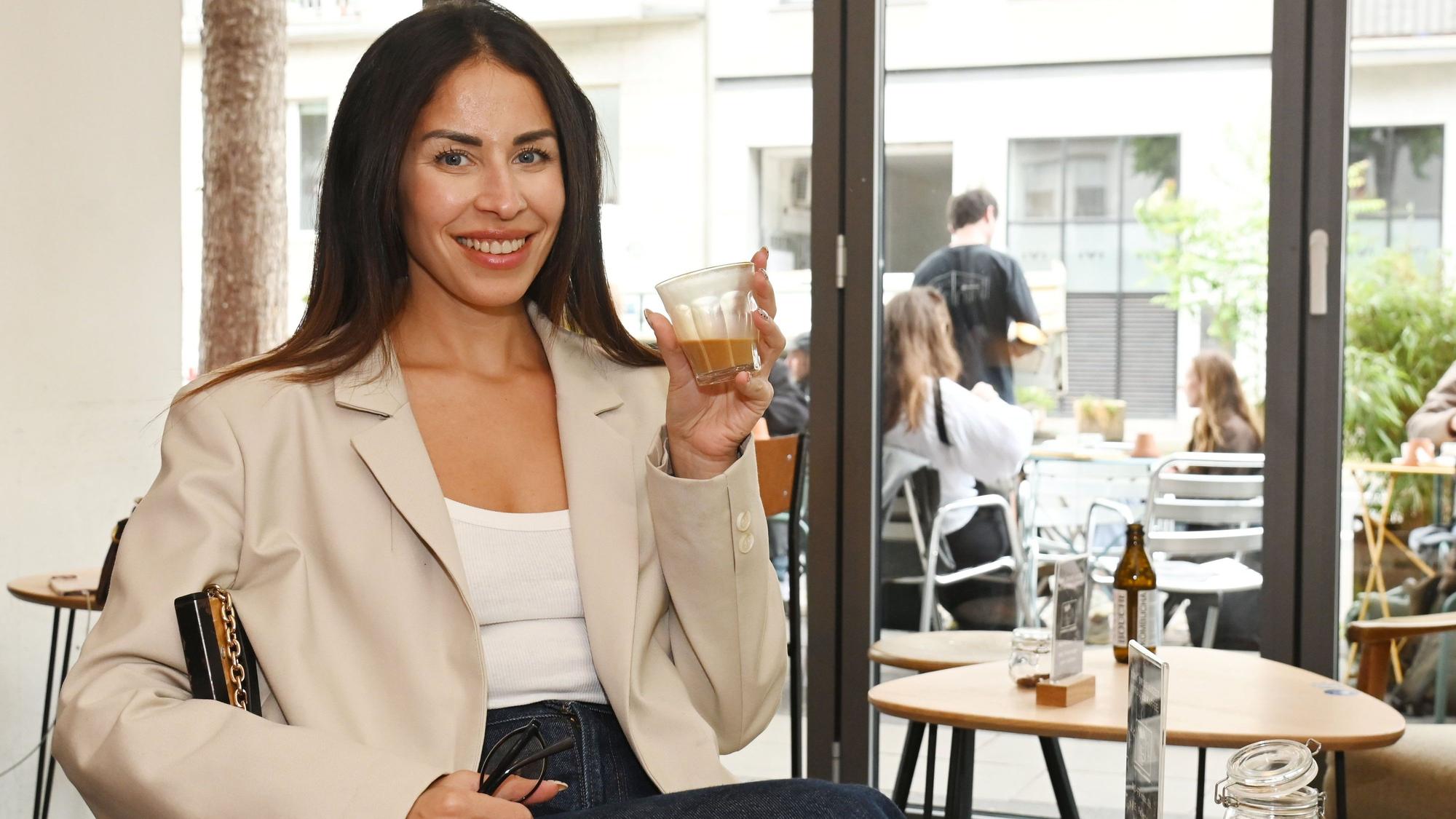 Eine junge Frau mit dunklem, langem Haar sitzt an einem Café-Tisch und hält ein kleines Glas Kaffee in der Hand.