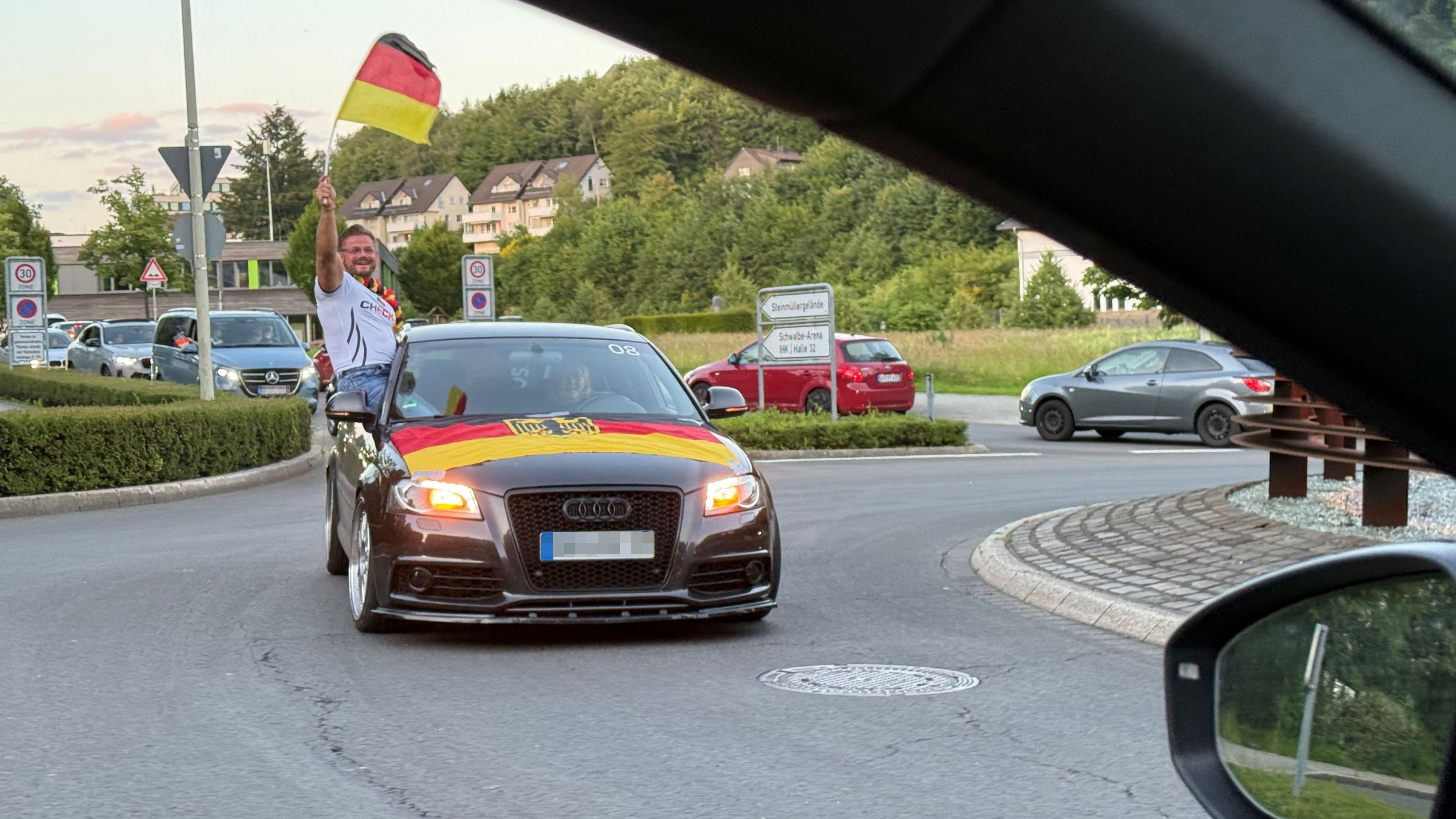 Das Foto zeigt einen Pkw. Über die Kühlerhaube ist die schwarz-rot-goldene Fahne mit Bundesadler gespannt. Der Beifahrer schwenkt die deutsche Fahne aus dem Fenster.