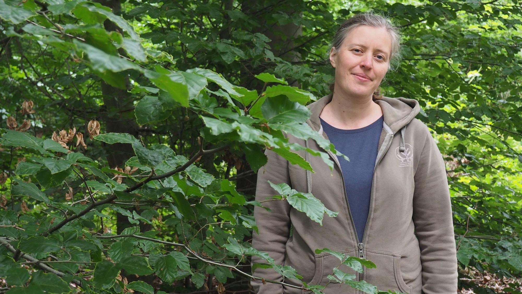 Katinka Richter trägt ein blaues Shirt und eine Kapuzenjacke und steht in einem Wald.