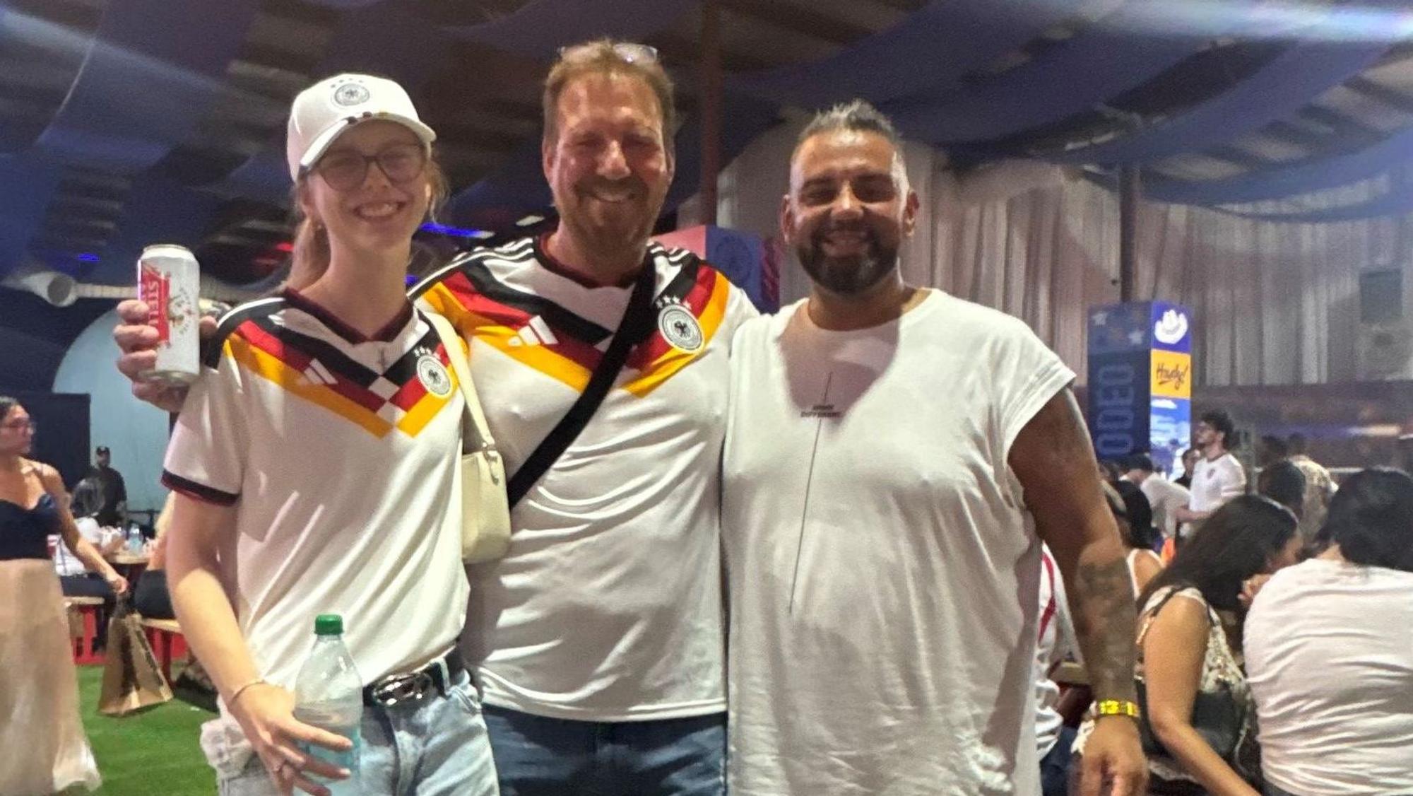Marcello Volk steht neben zwei Deutschland-Fans im Trikot.