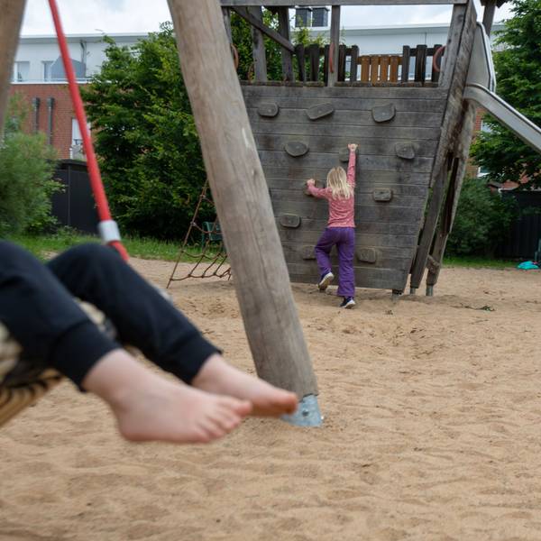 Auf einem Spielplatz ist ein schaukelndes Kind zu sehen. Im Hintergrund klettert ein Mädchen.