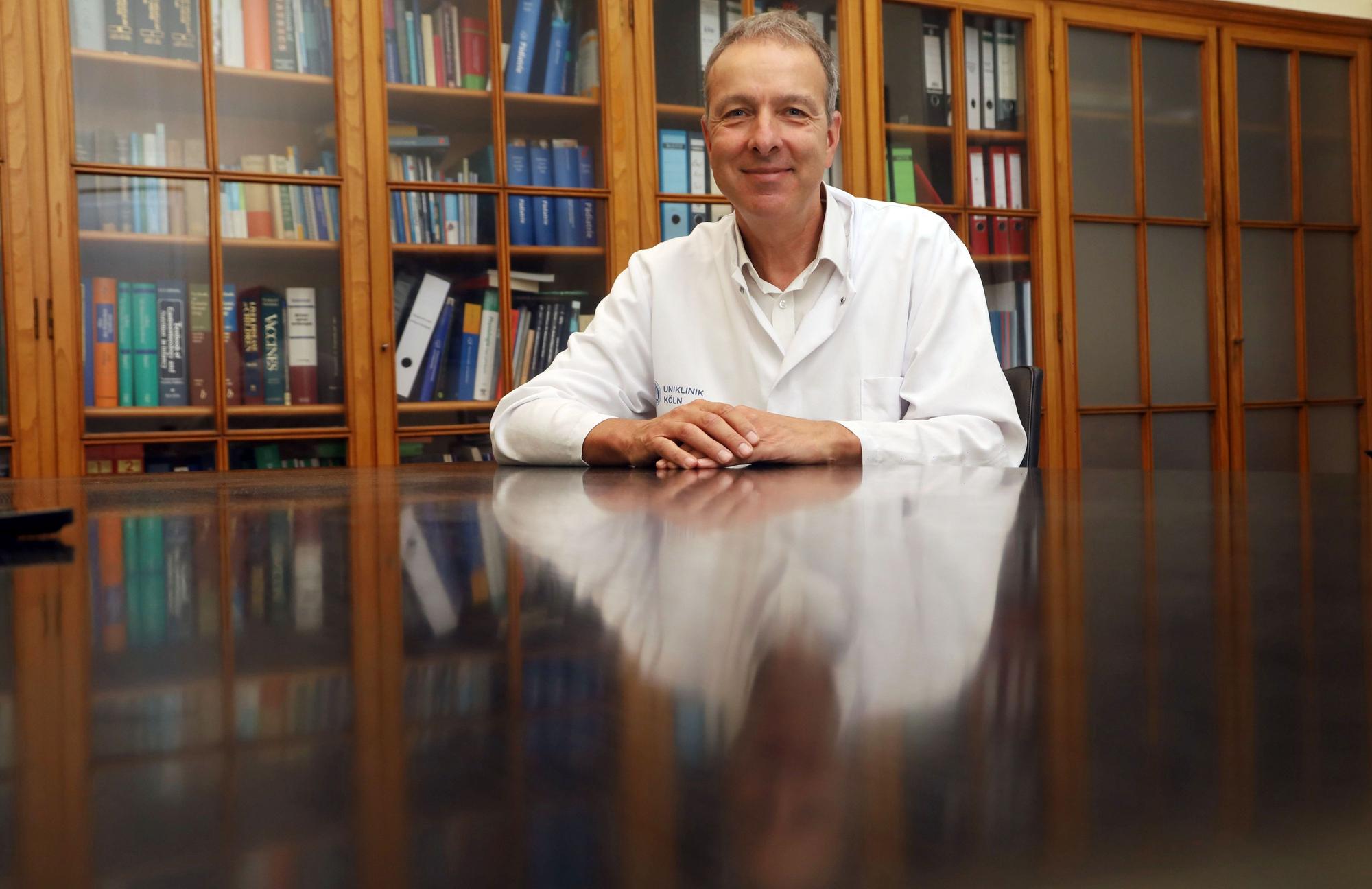 Professor Jörg Dötsch sitzt an einem Tisch, er trägt Arztkittel und lächelt in die Kamera.