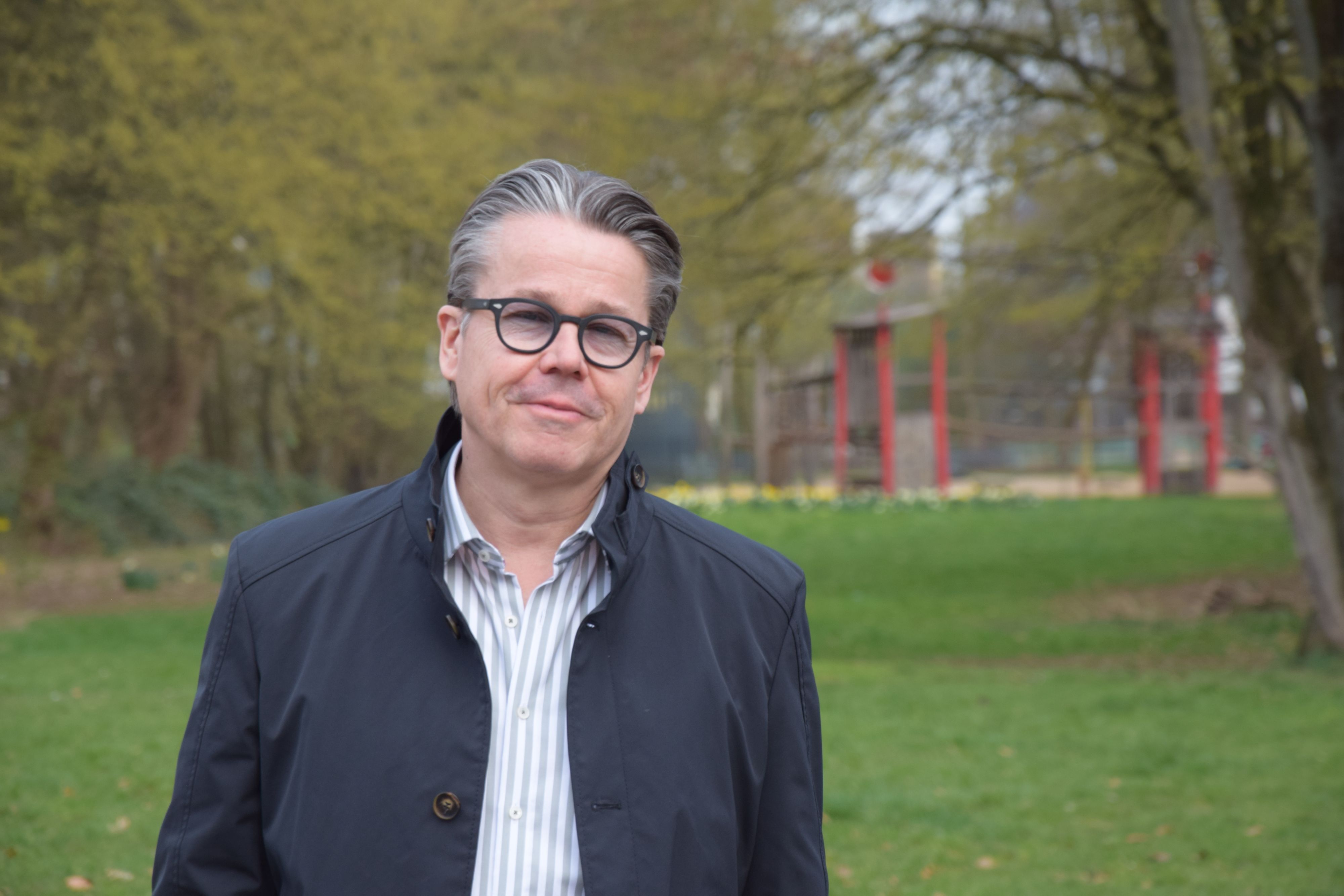 Ein Mann mit grauem Haar und Brille steht auf einer Rasenfläche. Im Hintergrund liegt ein Spielplatz.