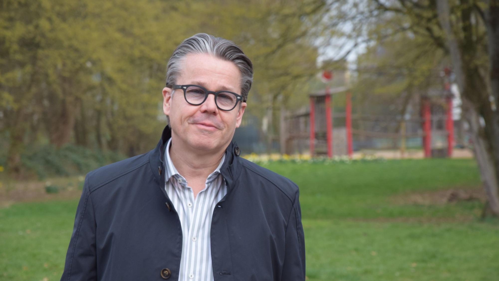 Ein Mann mit grauem Haar und Brille steht auf einer Rasenfläche. Im Hintergrund liegt ein Spielplatz.