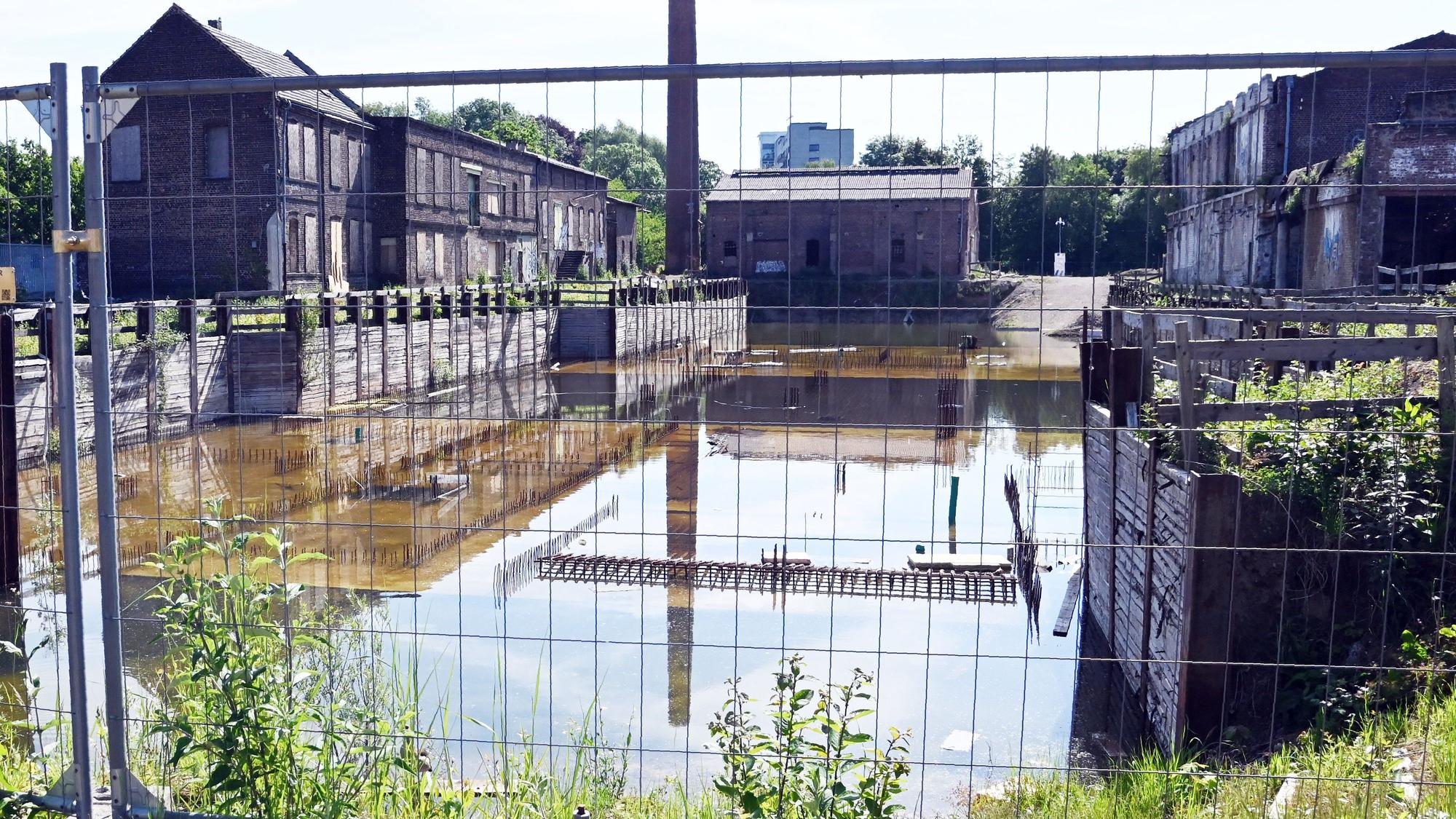 Ein altes Industriegelände mit Backstein-Fabrikgebäuden und Schornstein liegt hinter einem Bauzaun.