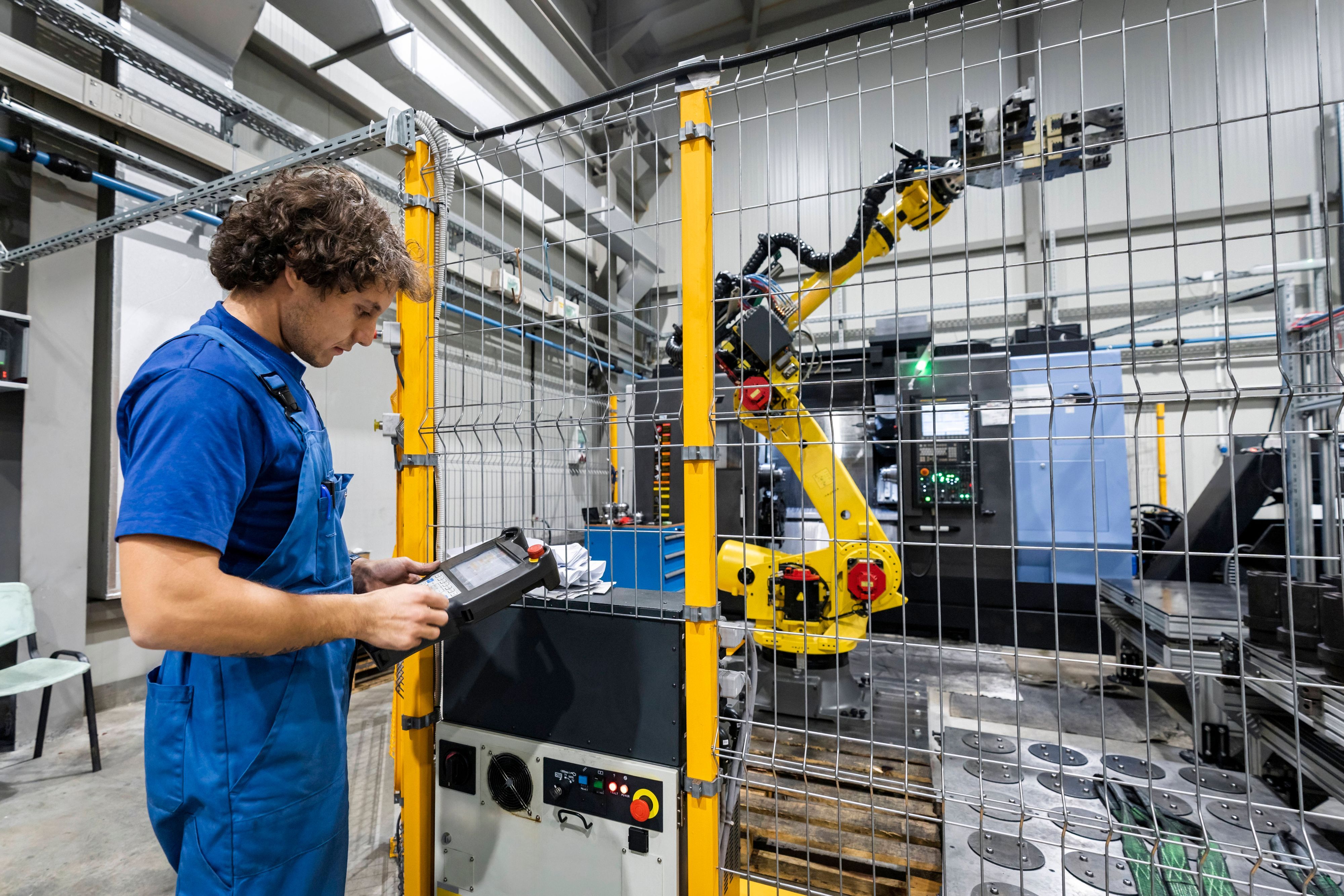 Ein Mann im Blaumann steht in einer Produktionshalle an einer Maschine mit Roboterarm.
