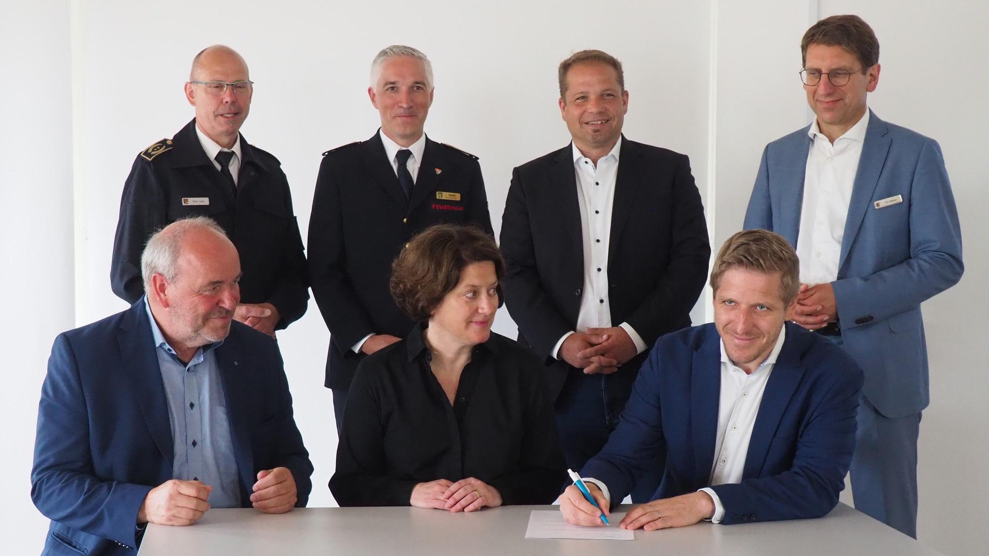 Zwei Männer und eine Frau sitzen an einem Tisch, einer unterschreibt ein Papier. Dahinter stehen vier Männer.