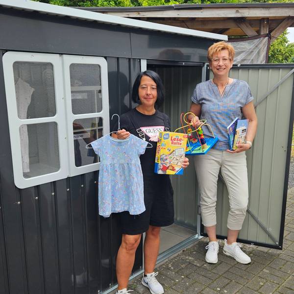 Zu sehen sind zwei Frauen mit Spielzeug und Kleidung für Kinder vor einem Gartenhäuschen.
