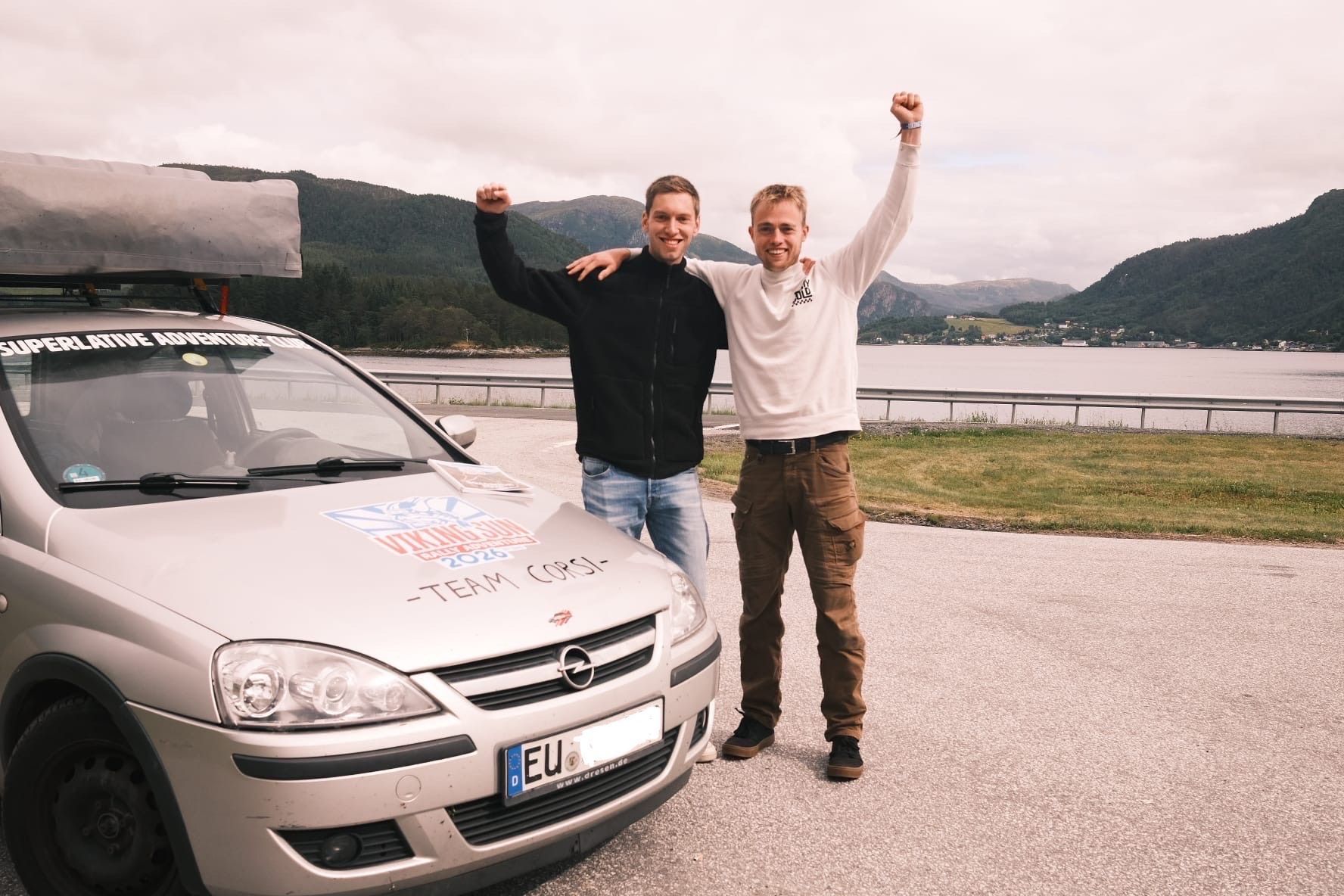 Die beiden jungen Männer stehen neben ihrem Opel Corsa, im Hintergrund ein See und  Berge.