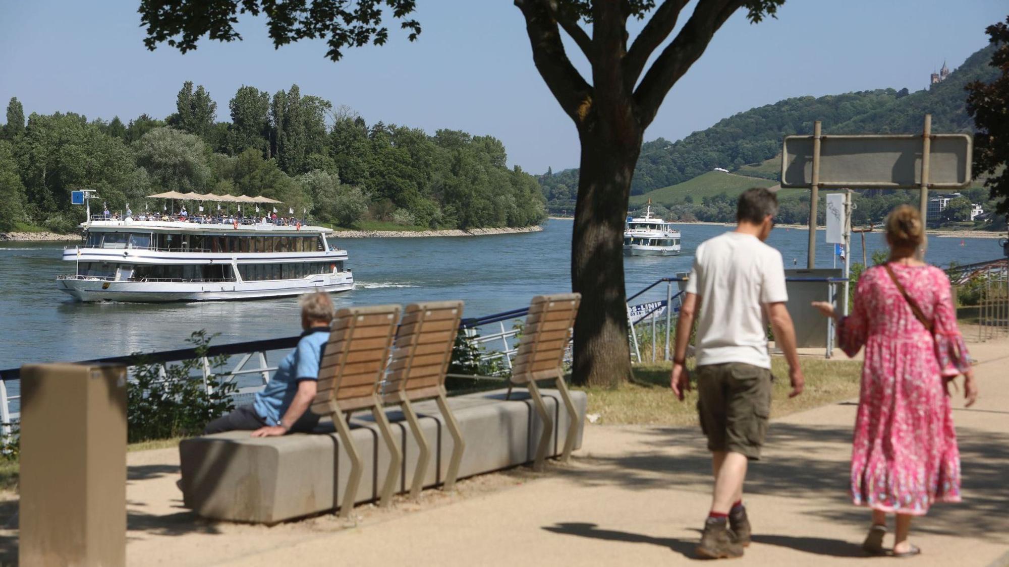 Ein Paar geht über die Insel, links sitzt eine Person auf einer Bank und blickt auf ein vorbeifahrendes Ausflugsschiff.