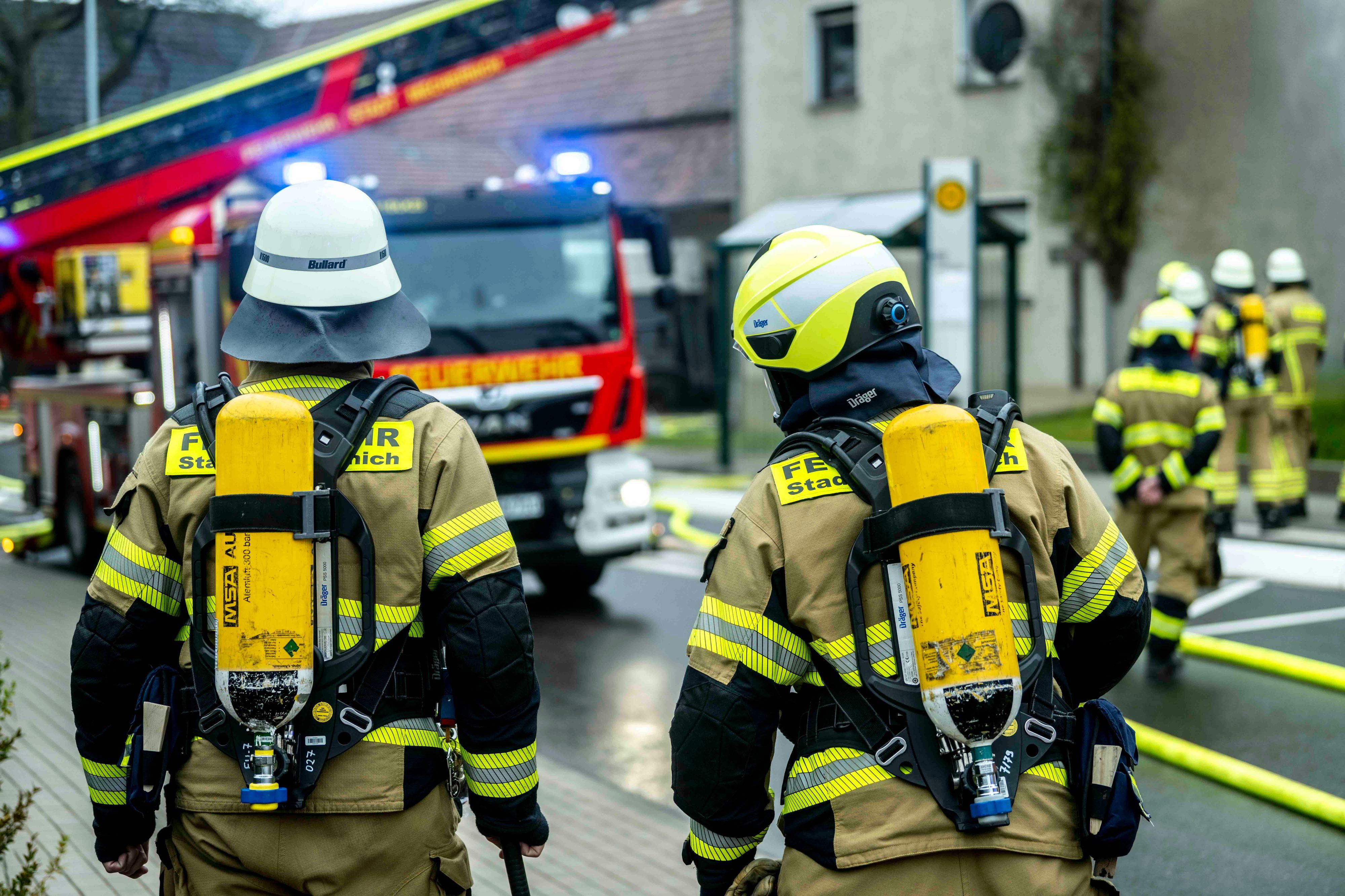 Zwei Feuerleute mit Pressluftatmern auf dem Rücken schauen auf eine Einsatzstelle, an der auch eine Drehleiter steht.