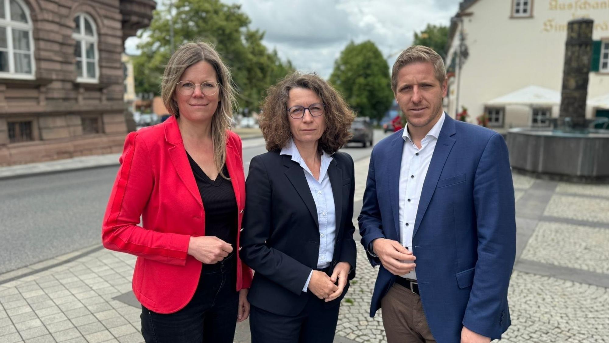 Die drei Obengenannten stehen auf einem gepflasterten Platz.