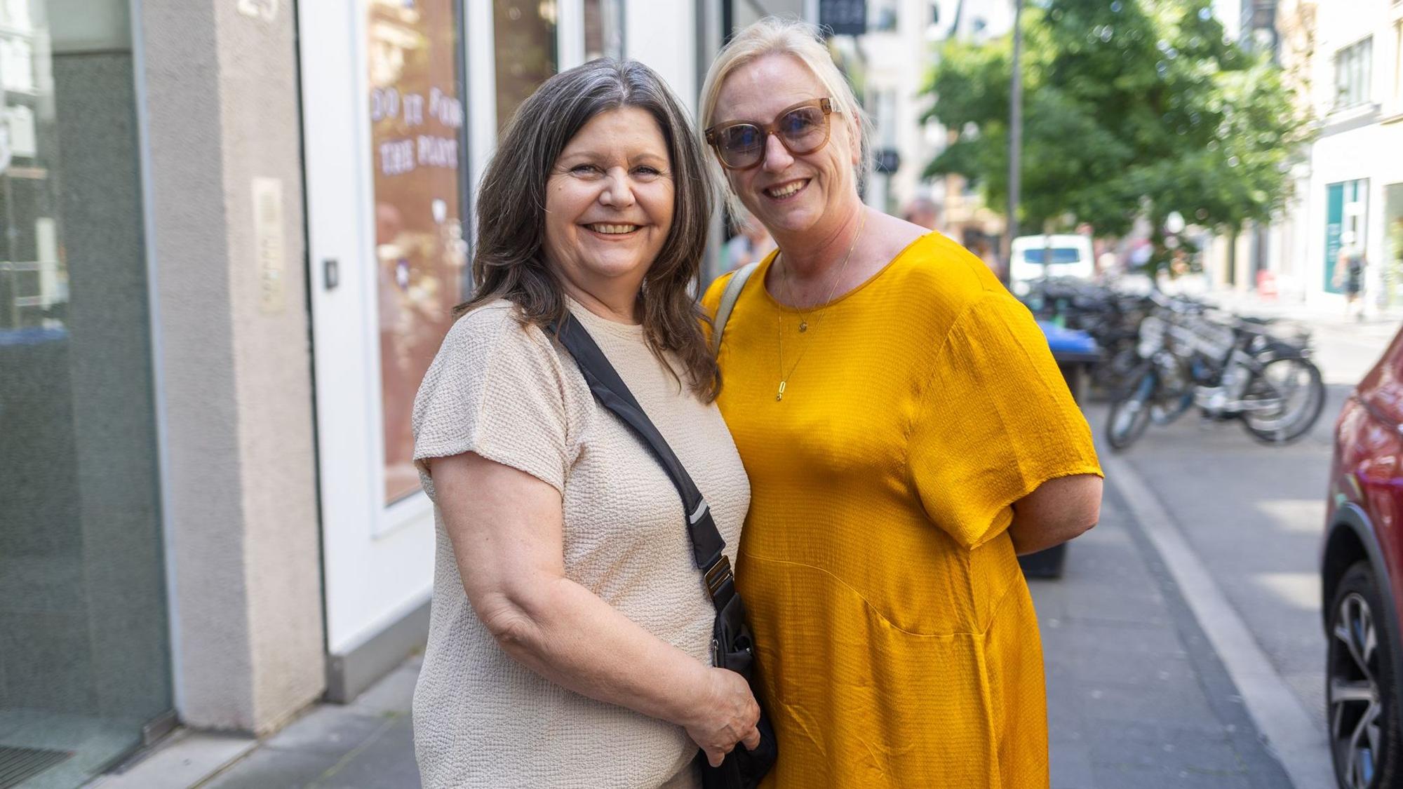 Zwei Frauen im braunen und gelben Kleid lächeln in die Kamera.