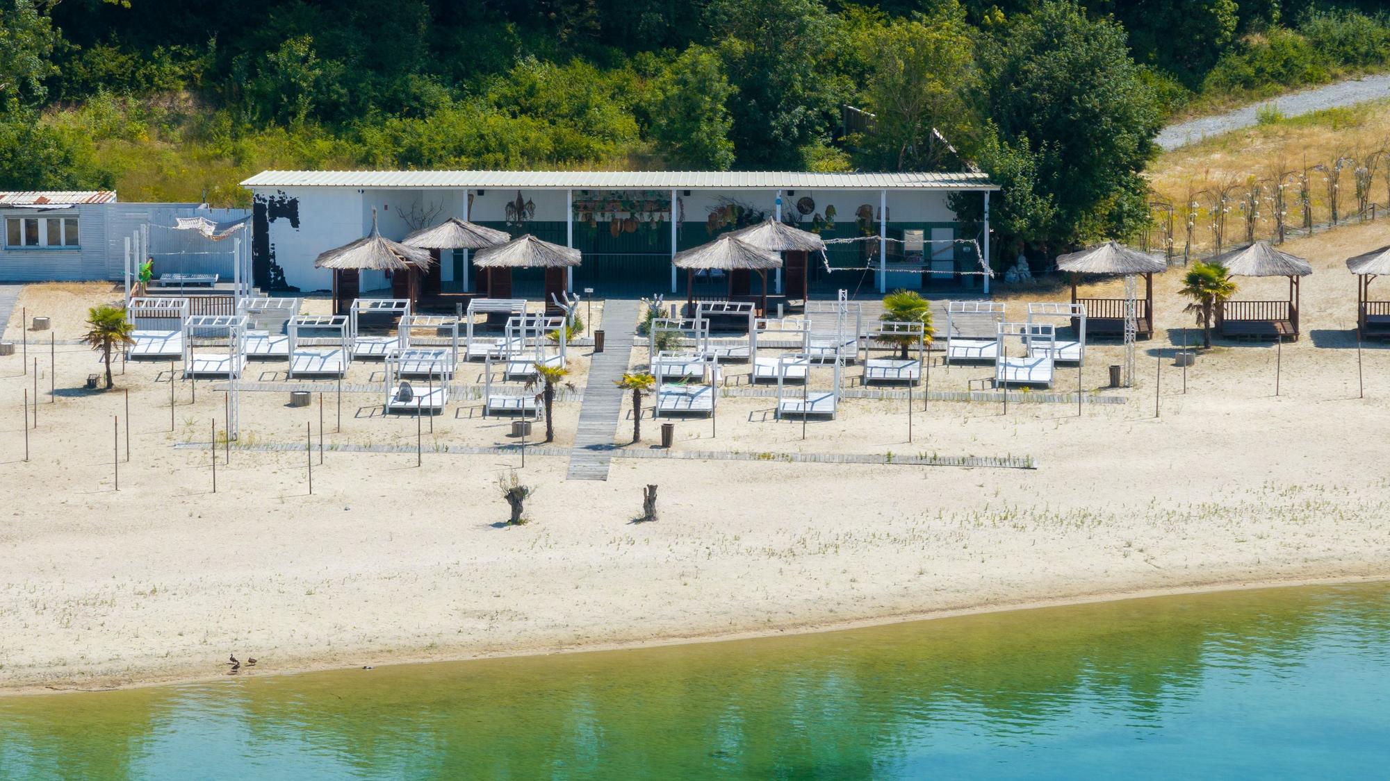 Am Strand eines Sees sind eine Beachbar, Sonnenhüte und Sonnenliegen. Die Anlage ist verwaist.