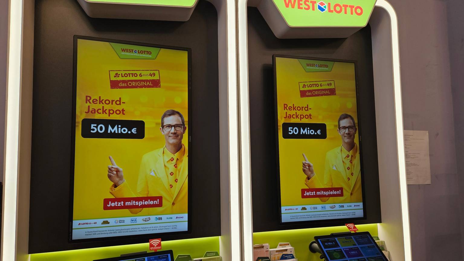 Auf dem Bild sieht man zwei WESTLOTTO-Terminals in einer Lotto-Annahmestelle mit großen Werbedisplays. Auf den Bildschirmen wird ein Rekord-Jackpot von 50 Millionen Euro für LOTTO 6aus49 beworben.