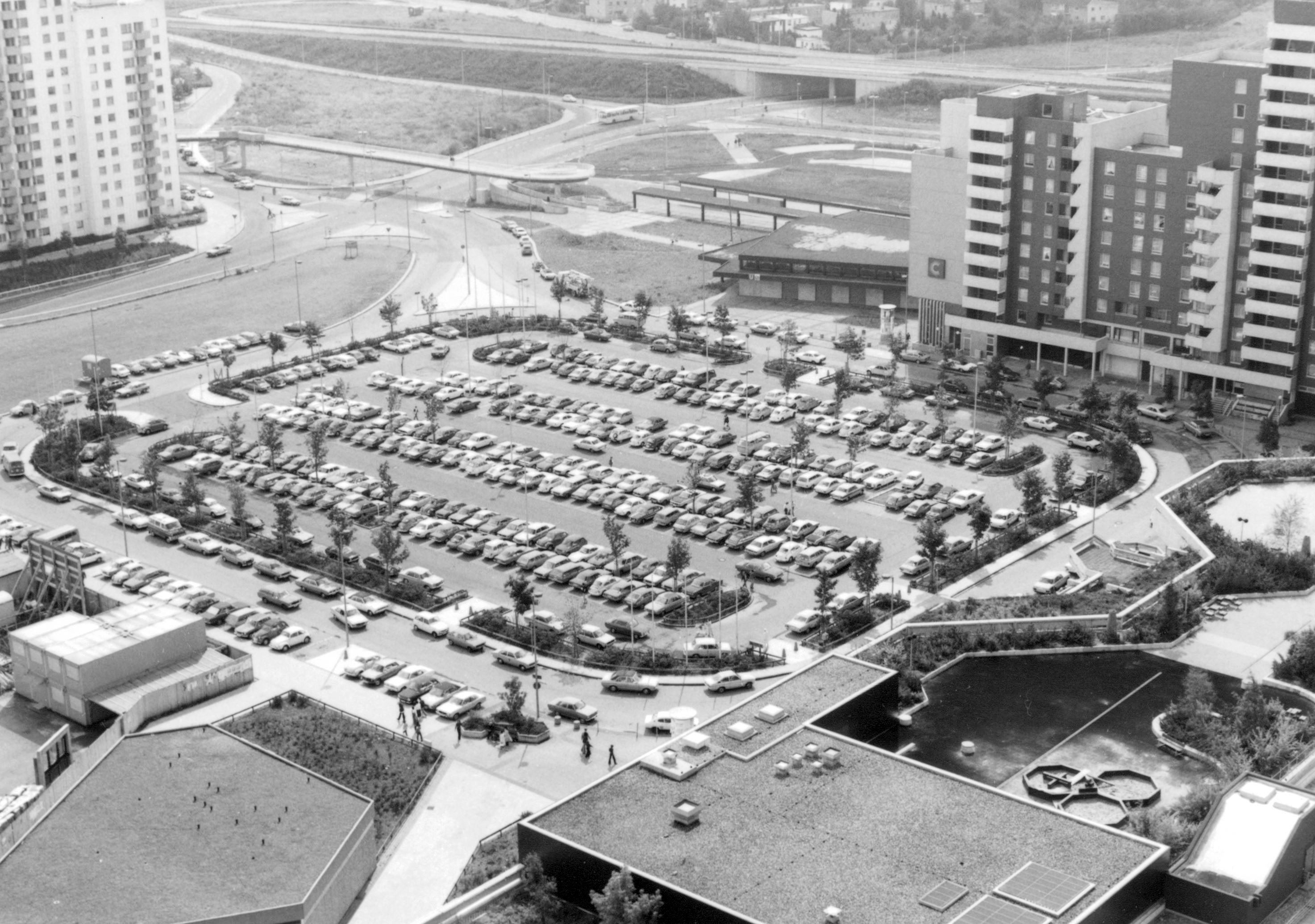 Eine Schwarz-Weiß-Luftaufnahme zeigt einen riesigen Parkplatz, an den sich das Gebäude eines Einkaufszentrums anschließt. Außerdem sind Hochhäuser zu sehen.