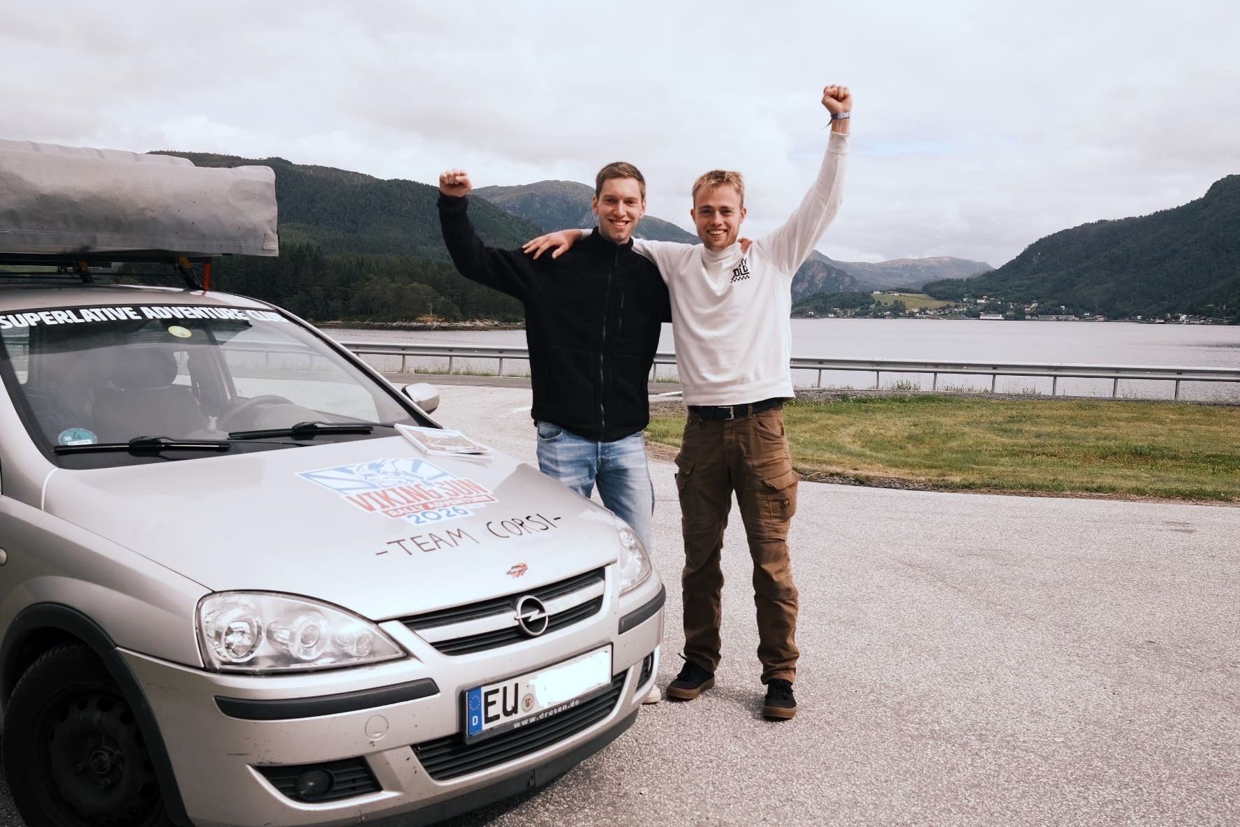 Die Zülpicher Henrik Krewel (l.) und Andreas Ulm stehen neben ihrem Opel Corsa und recken jubelnd je einen Arm in die Höhe. Im Hintergrund ist ein See zu sehen.