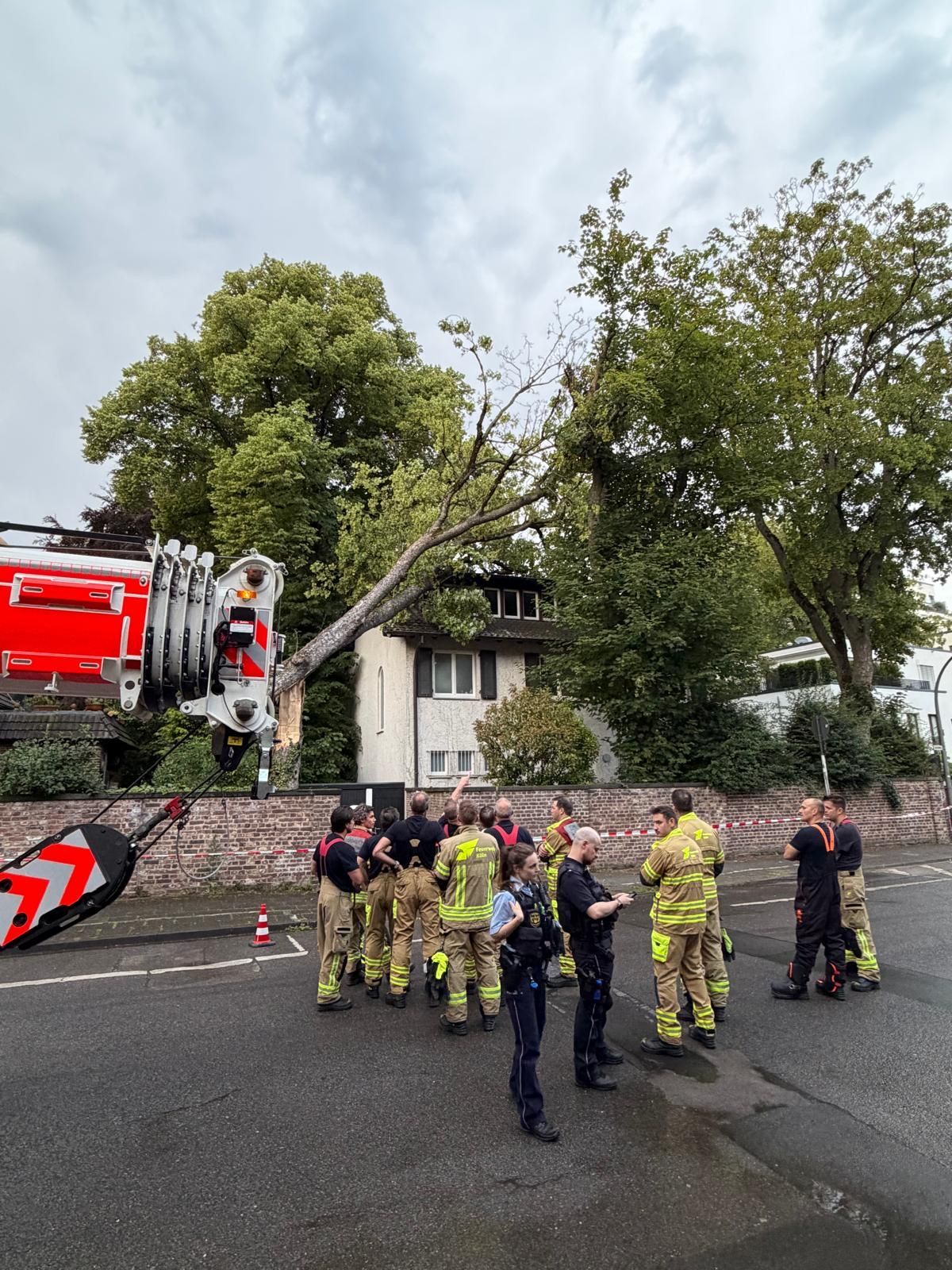 Ein Baum ist auf ein Haus gekippt, davor stehen zahlreiche Einsatzkräfte von Feuerwehr und Polizei.