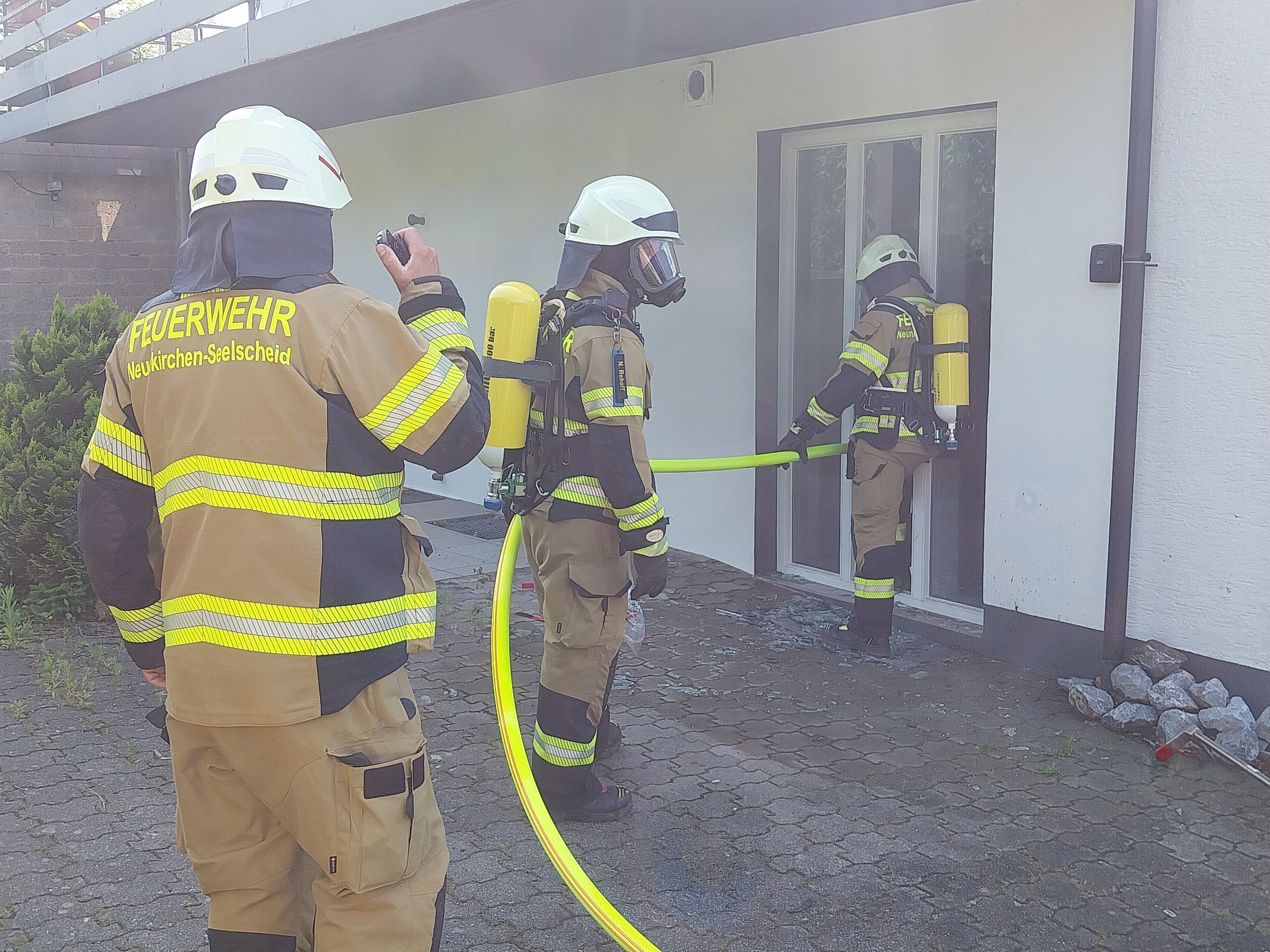 Drei Feuerwehrleute stehen mit einem Löschschlauch vor dem Erdgeschoss eines Hauses.