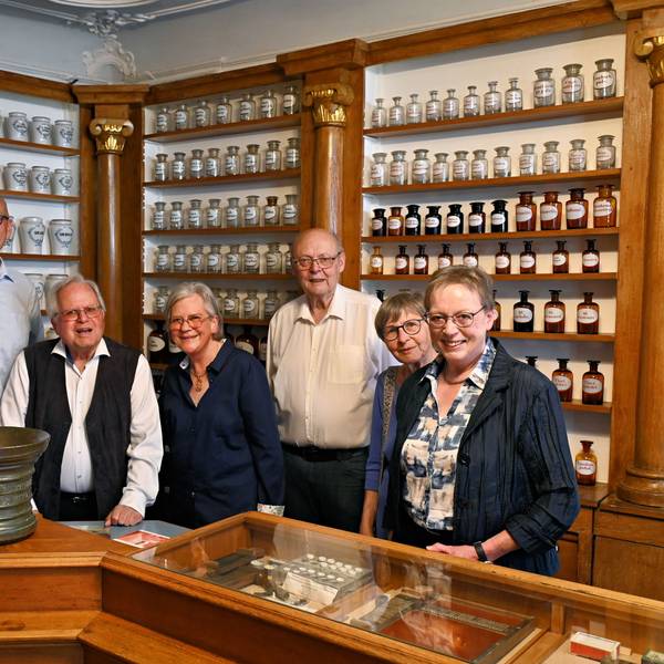 Die genannten Personen stehen im historischen Apothekengebäude vor Regalen mit zahlreichen Flaschen und anderen Behältnissen.