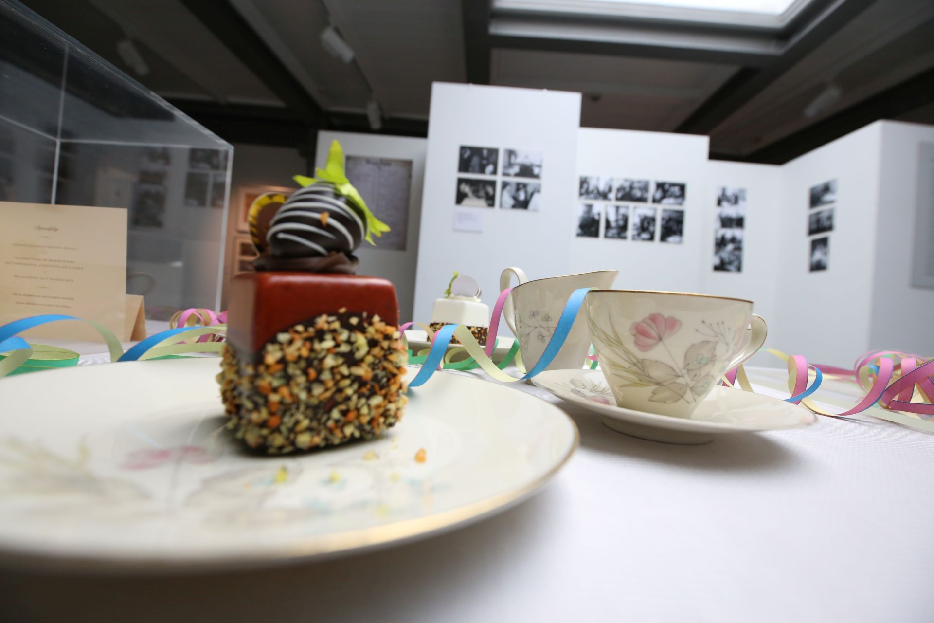 Eine Kaffeetafel mit Kuchen, Tellern und Tassen; im Hintergrund Fotos an einer Stellwand.