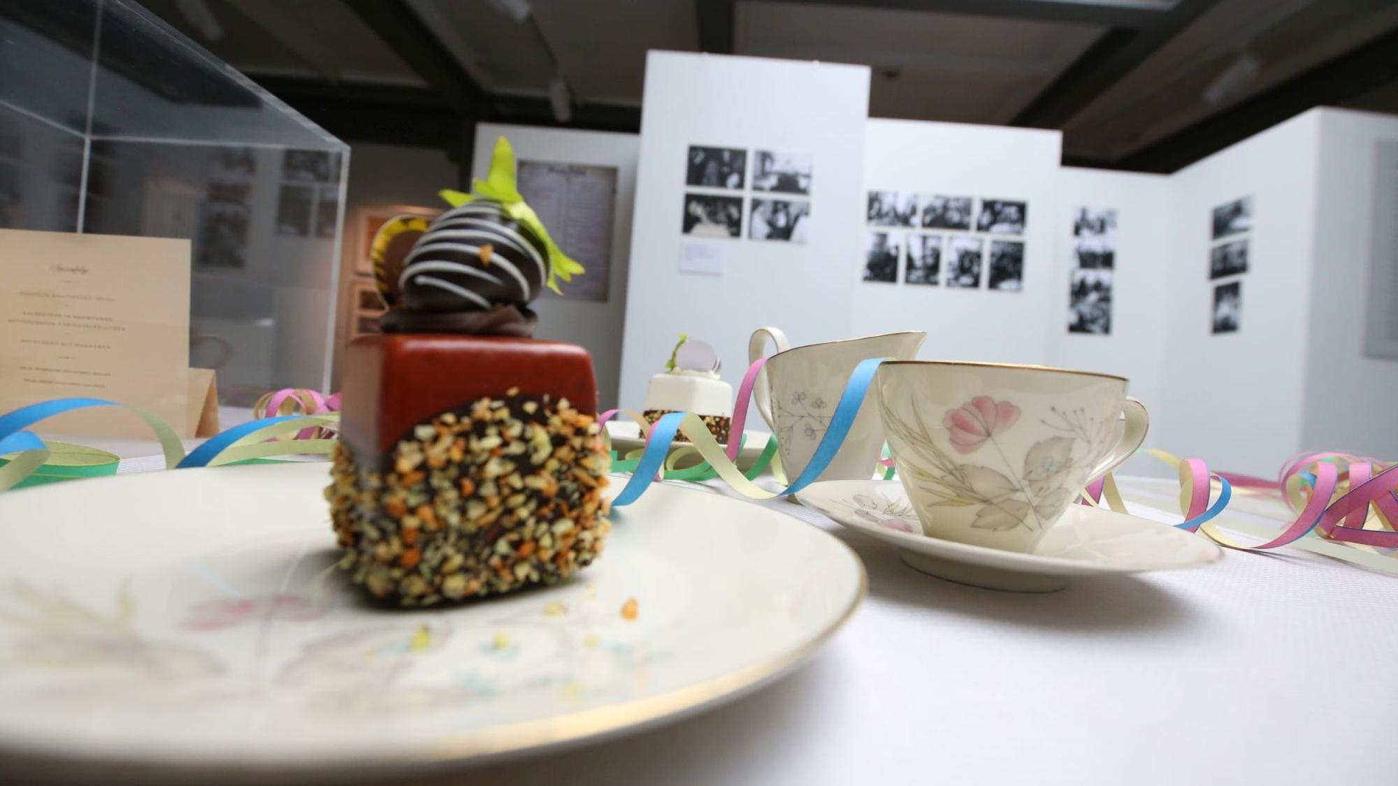 Eine Kaffeetafel mit Kuchen, Tellern und Tassen; im Hintergrund Fotos an einer Stellwand.