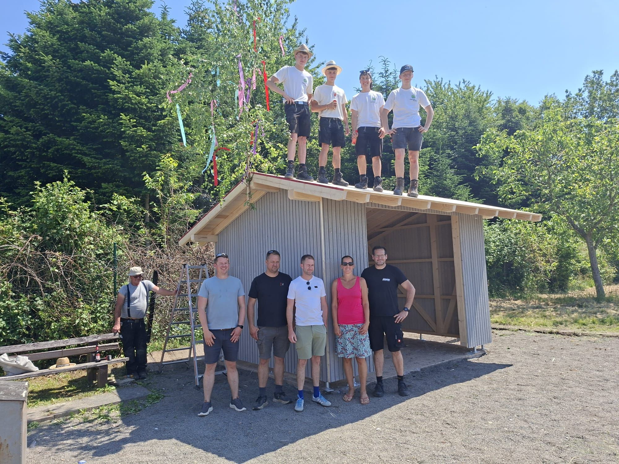 Zu sehen sind einige Menschen vor einer nicht ganz fertig gebauten Hütte im Grünen.