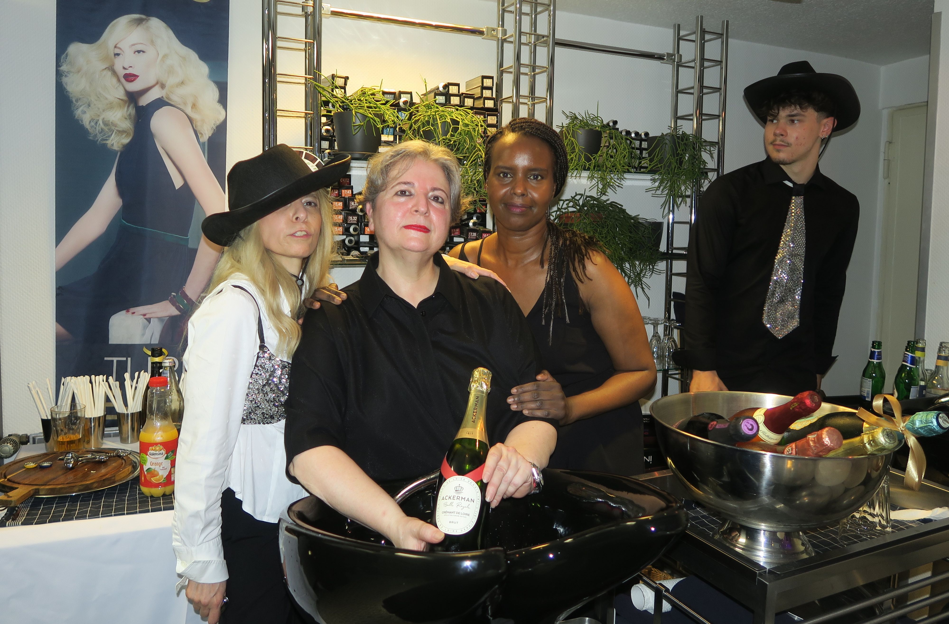 Drei Frauen in festlicher Kleidung stehen hinter einem Haarwaschbecken in einem Friseursalon. In einem großen Sektkühler liegen viele Flaschen kalt.