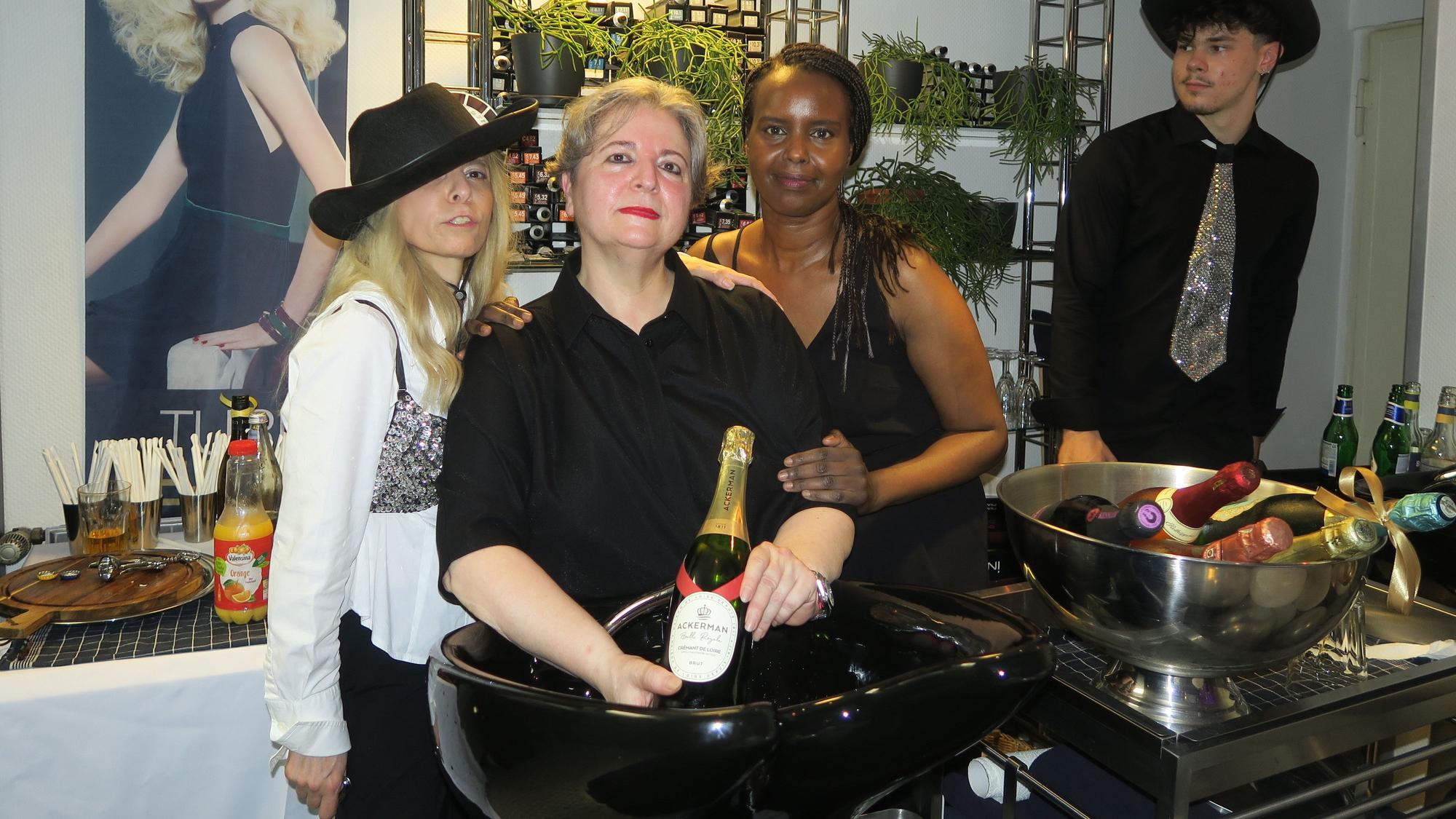 Drei Frauen in festlicher Kleidung stehen hinter einem Haarwaschbecken in einem Friseursalon. In einem großen Sektkühler liegen viele Flaschen kalt.