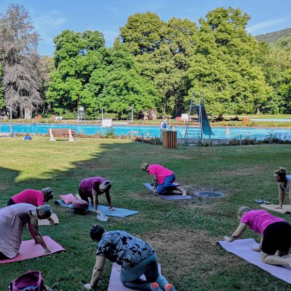Auf der Liegewiese des Gemünder Rosenbads nehmen mehrere Frauen an einer Yogastunde teil. Im Hintergrund ist das Schwimmbecken zu sehen.