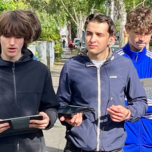 Zwei Schüler stehen auf einer Straße, zwei haben Tablets in den Händen, einer spricht.
