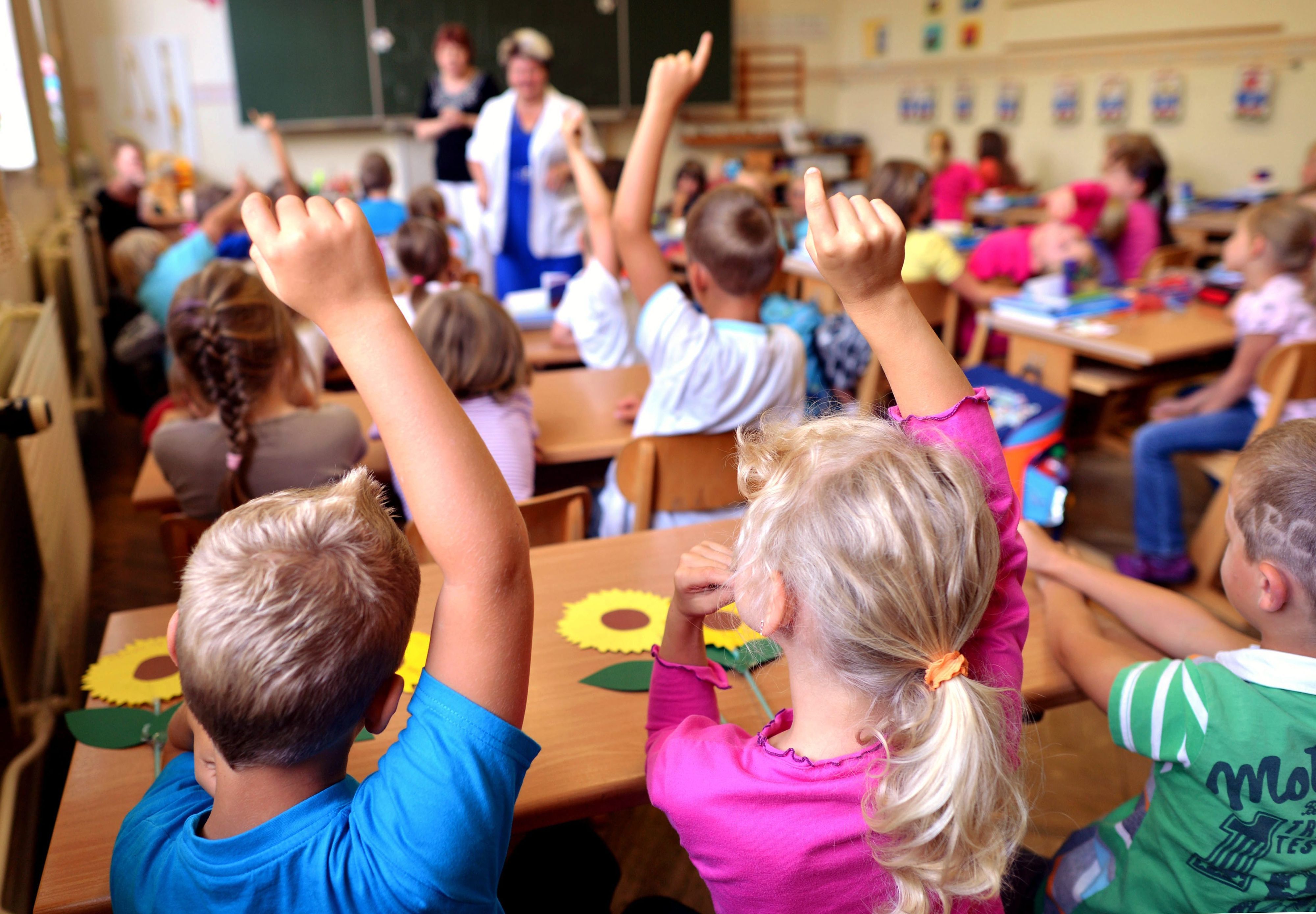 Kinder sitzen an Holztischen in einem Klassenraum.