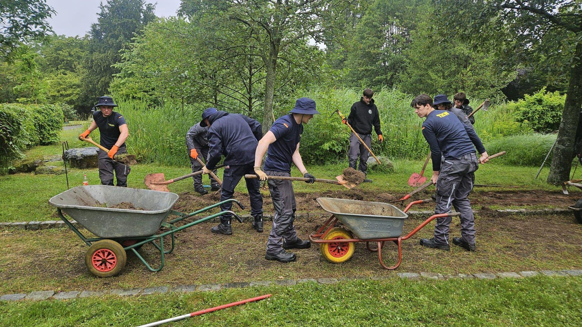 Mehrere Männer arbeiten mit Schaufeln in einer Grünanlage. Sie haben auch zwei Schubkarren dabei.