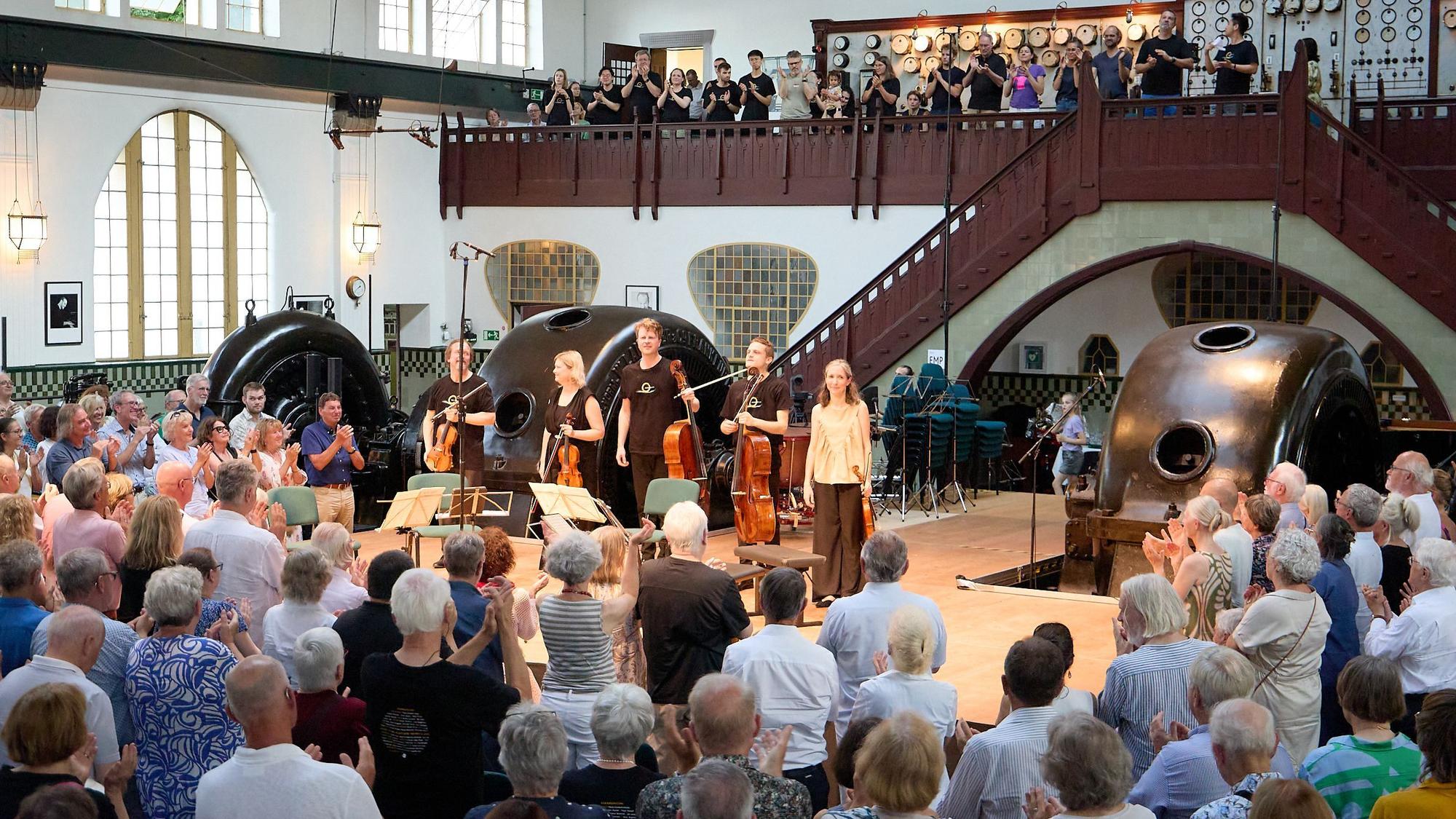Das Foto zeigt das Publikum, das stehend applaudiert, von hinten. Auf der Bühne sind die Musiker zu sehen.