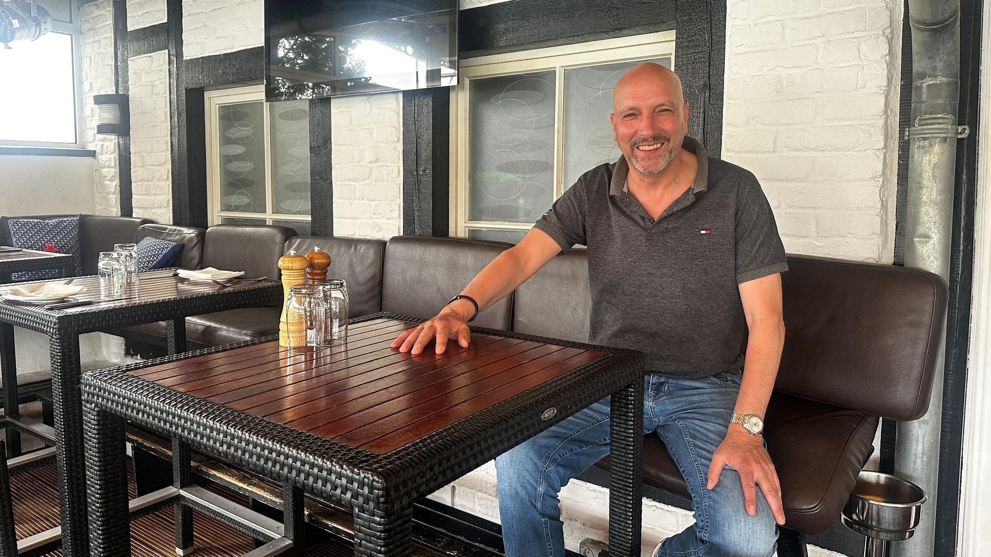 Ein Mann mit Glatze, Bart, Poloshirt und Jeans sitzt an einem Hochtisch.