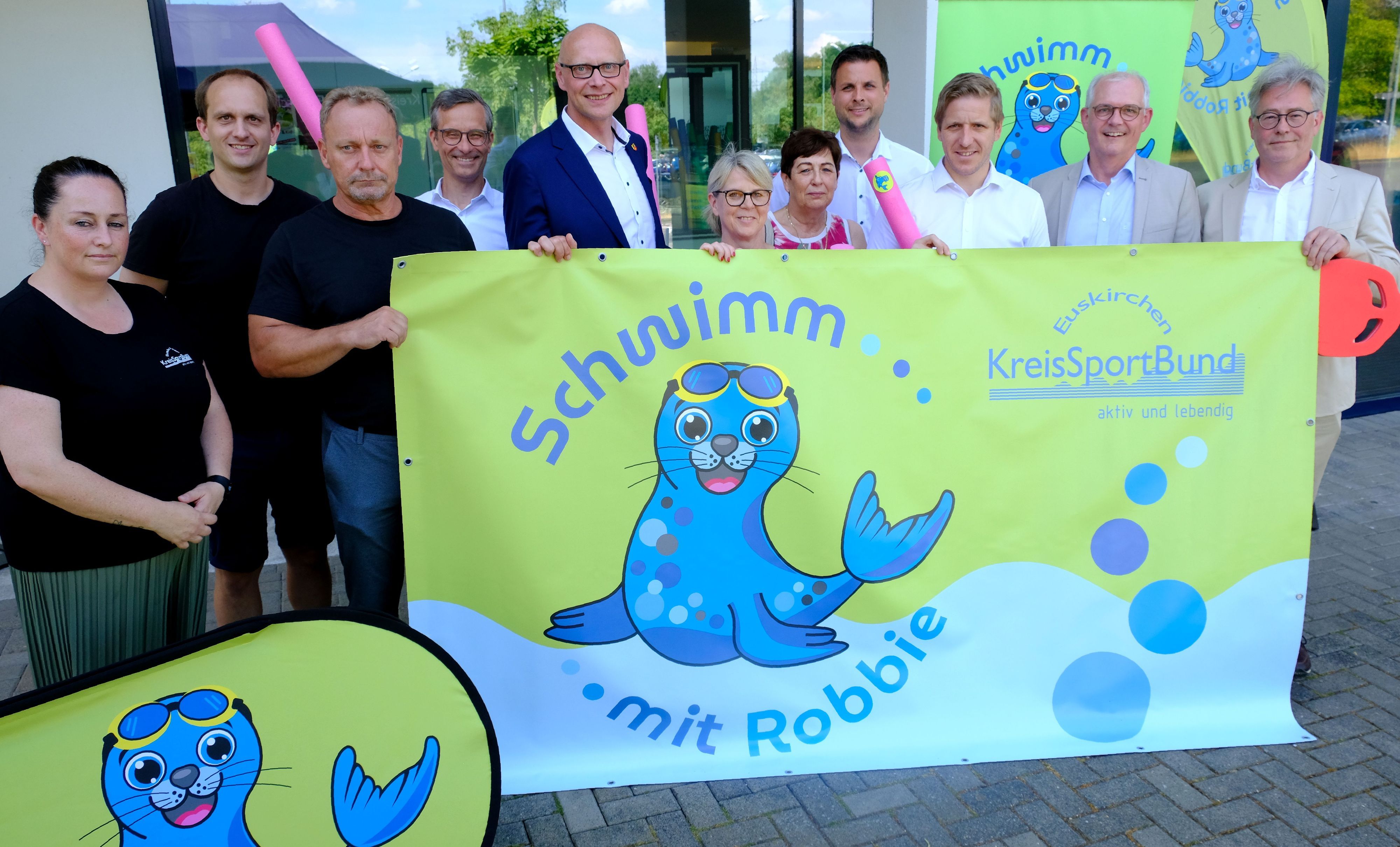 Vertreter der beteiligten Kommunen und des Kreissportbunds Euskirchen halten ein Banner mit der Aufschrift „Schwimm mit Robbie“ fest, auf dem eine Robbe im Wasser zu sehen ist.