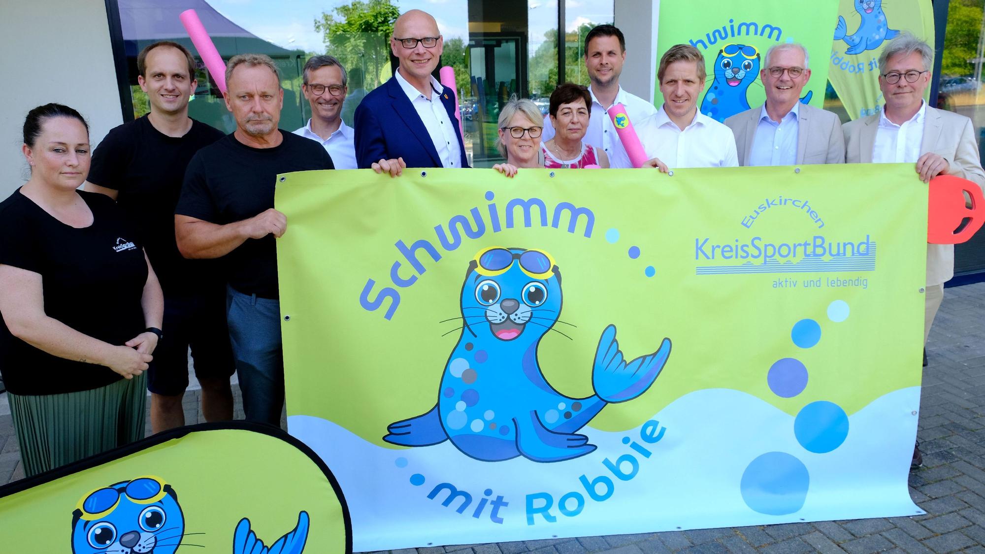 Vertreter der beteiligten Kommunen und des Kreissportbunds Euskirchen halten ein Banner mit der Aufschrift „Schwimm mit Robbie“ fest, auf dem eine Robbe im Wasser zu sehen ist.