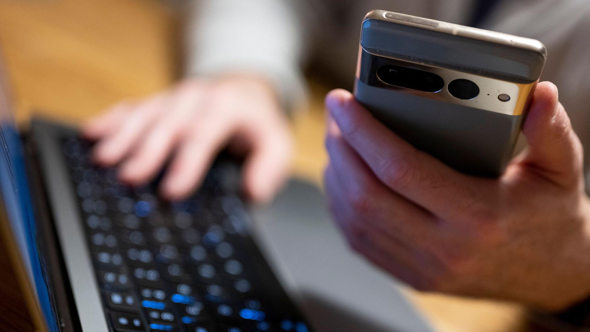 Ein Mann sitzt am Laptop und hält ein Smartphone in der Hand.