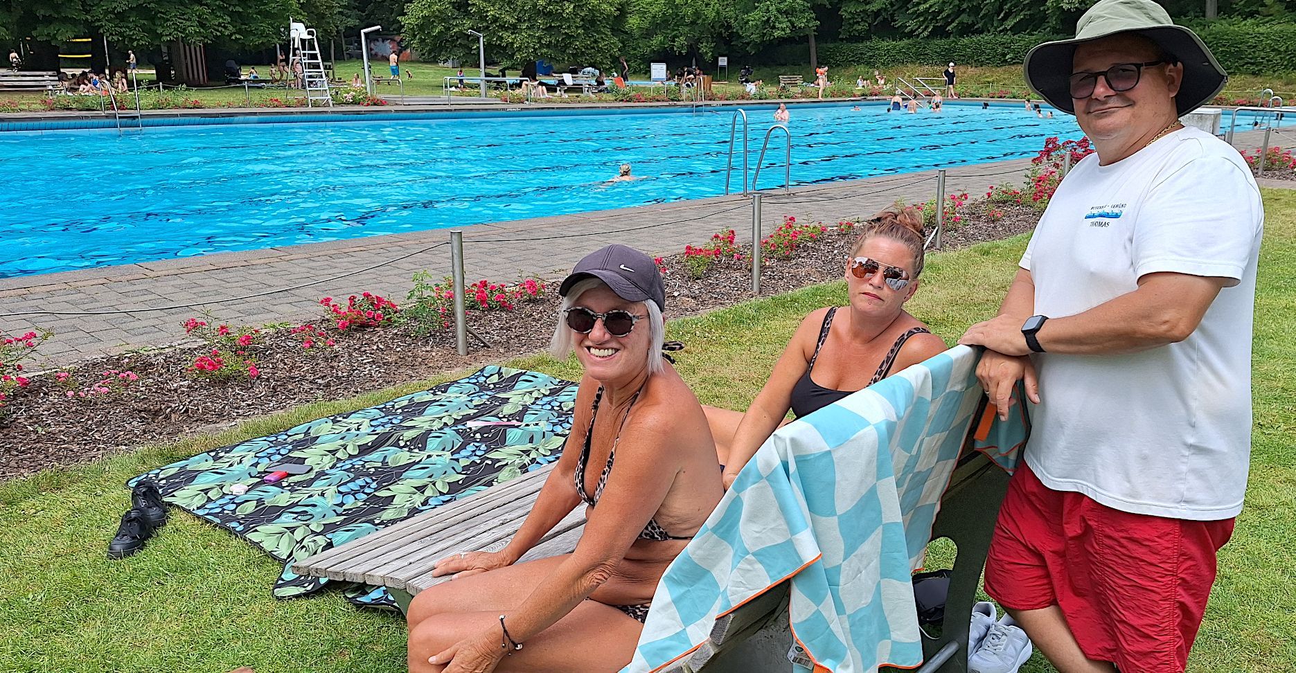 Zwei Frauen sitzen auf einer Liege, ein Mann steht daneben. Im Hintergrund ist das Schwimmbecken zu sehen.