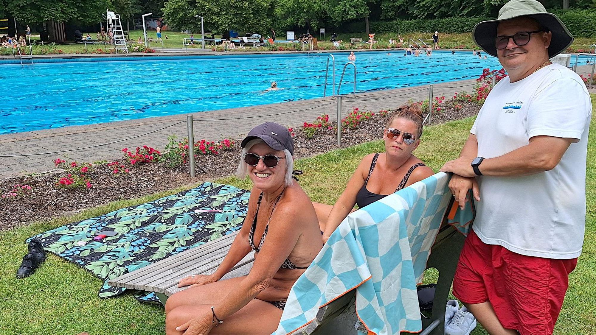 Zwei Frauen sitzen auf einer Liege, ein Mann steht daneben. Im Hintergrund ist das Schwimmbecken zu sehen.
