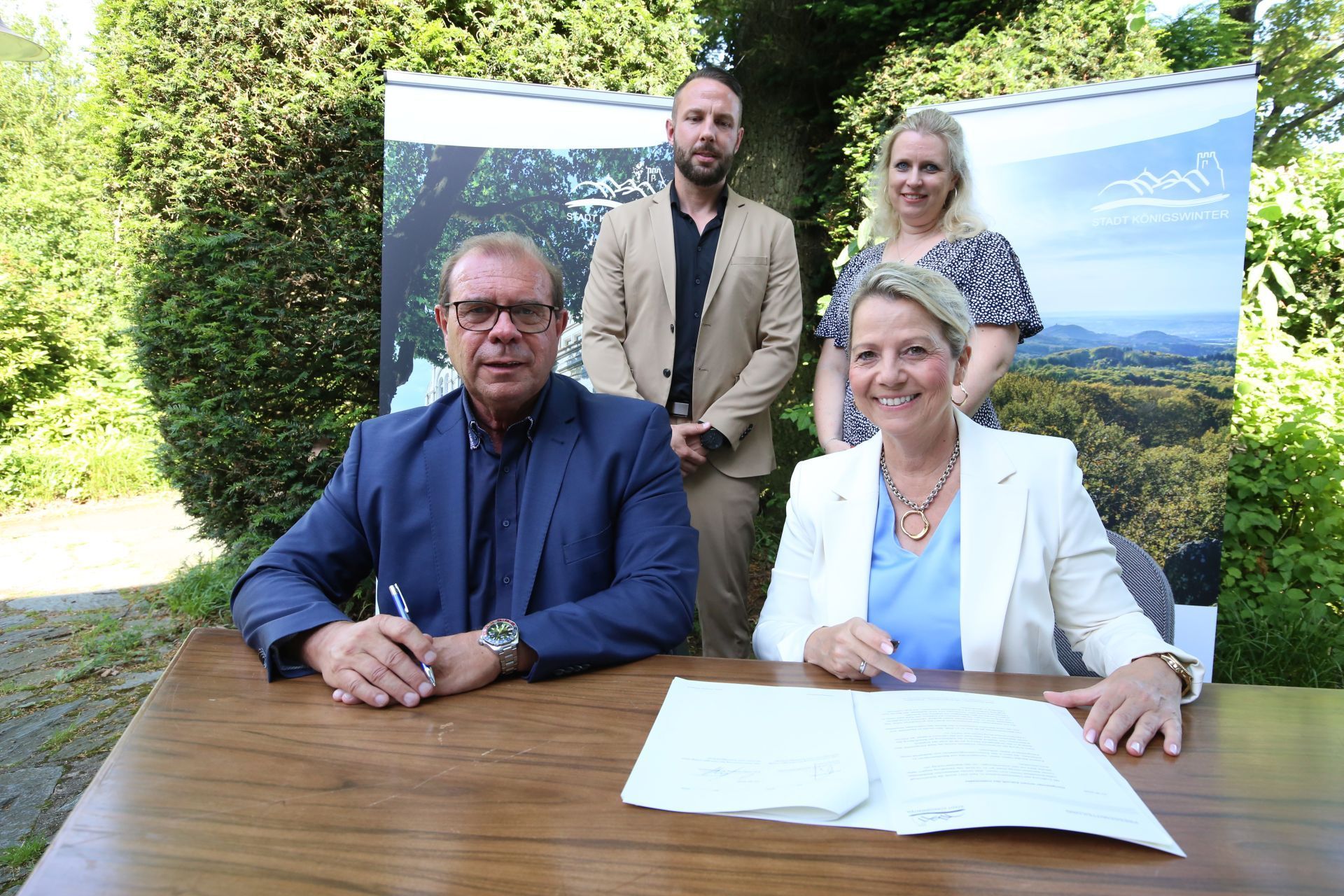 Vier Menschen; ein Mann und eine Frau sitzen an einem Tisch, auf dem ein Vertrag liegt.