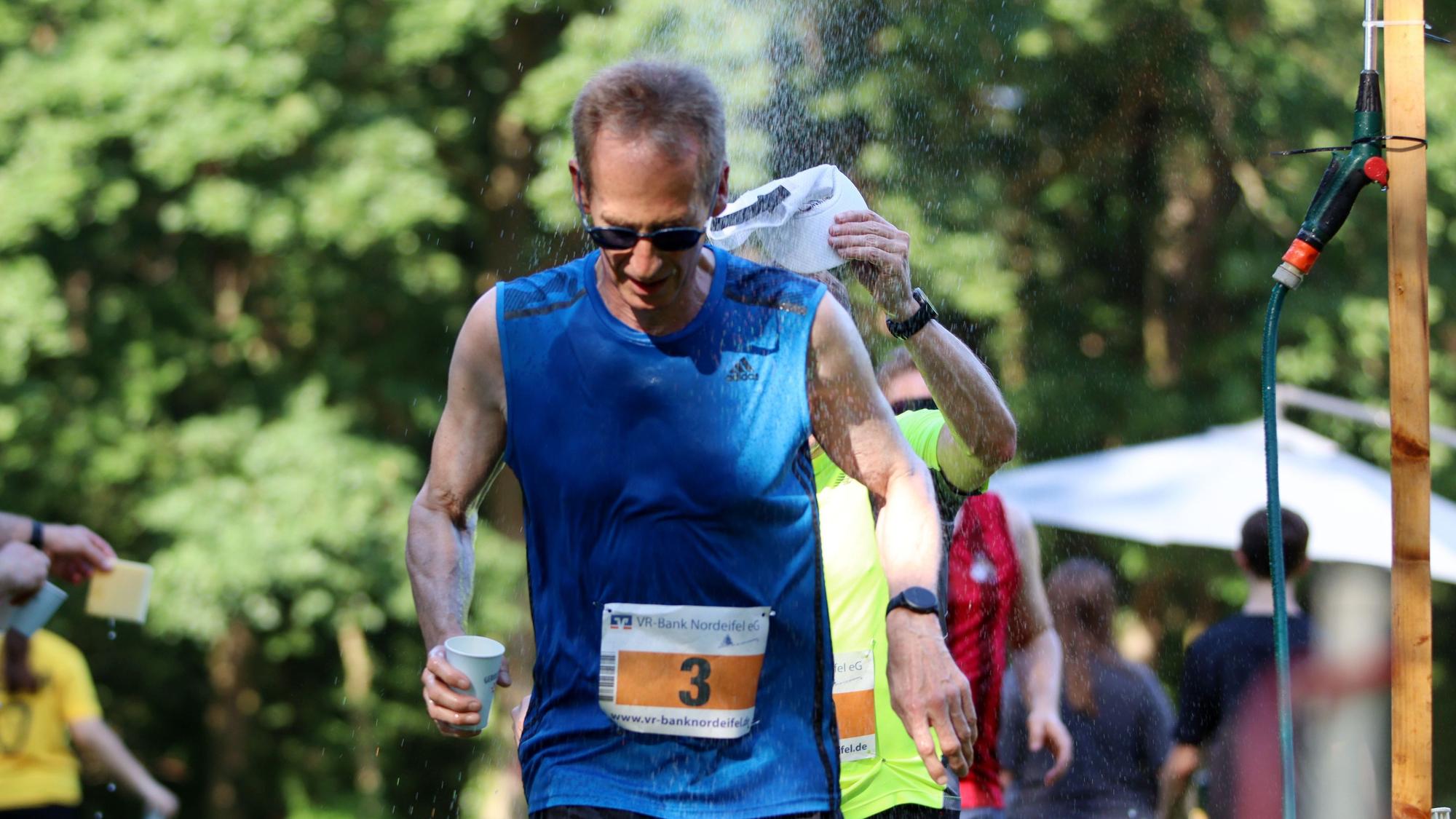 Detlef Seif läuft, mit einem Becher Wasser in der Hand, durch die Streckendusche.