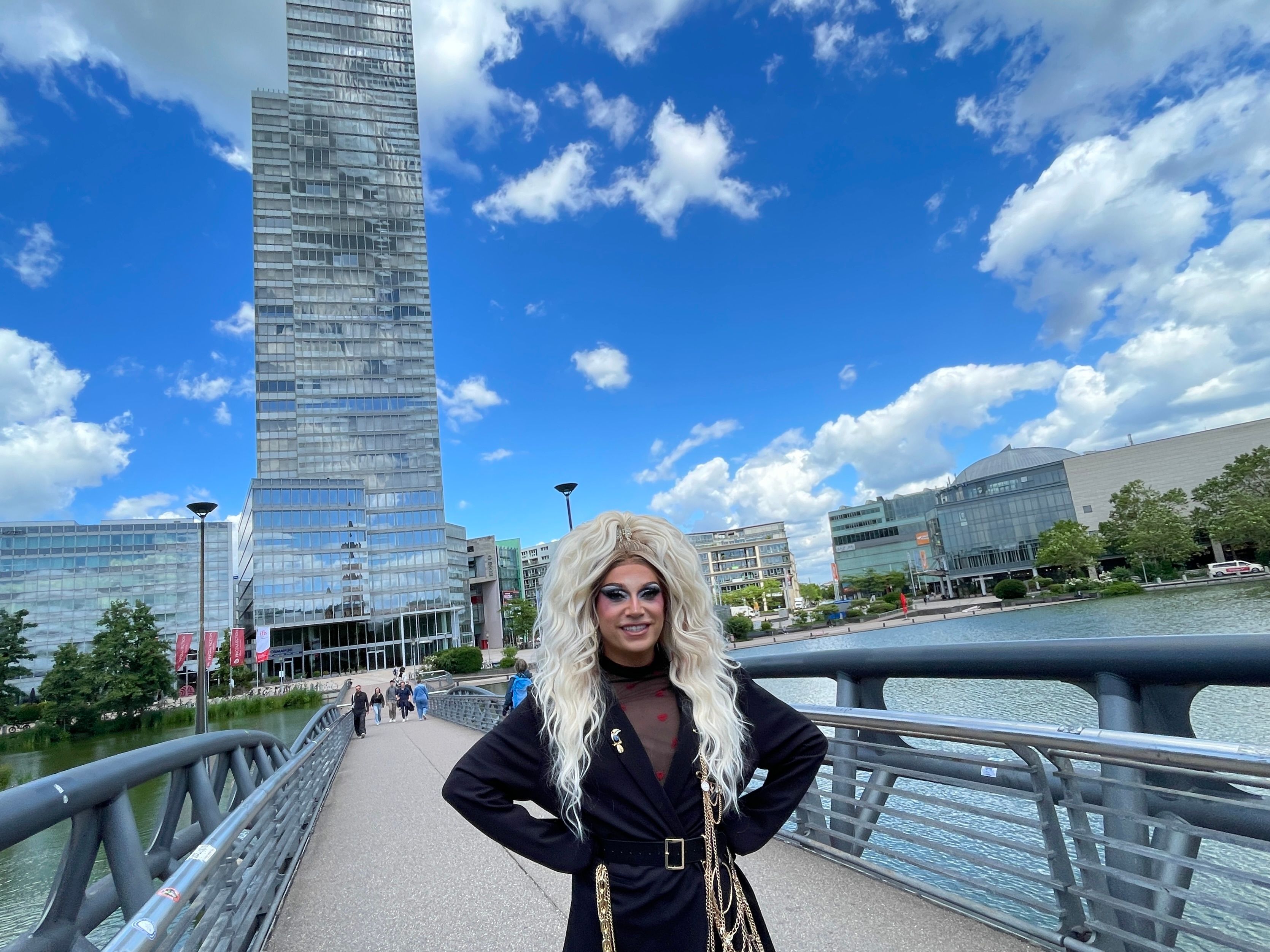 Eine Drag Queen mit blonder langer Perücke und schwarzem Mantel steht auf einer Brücke, im Hintergrund ist der Mediapark-Turm zu sehen.